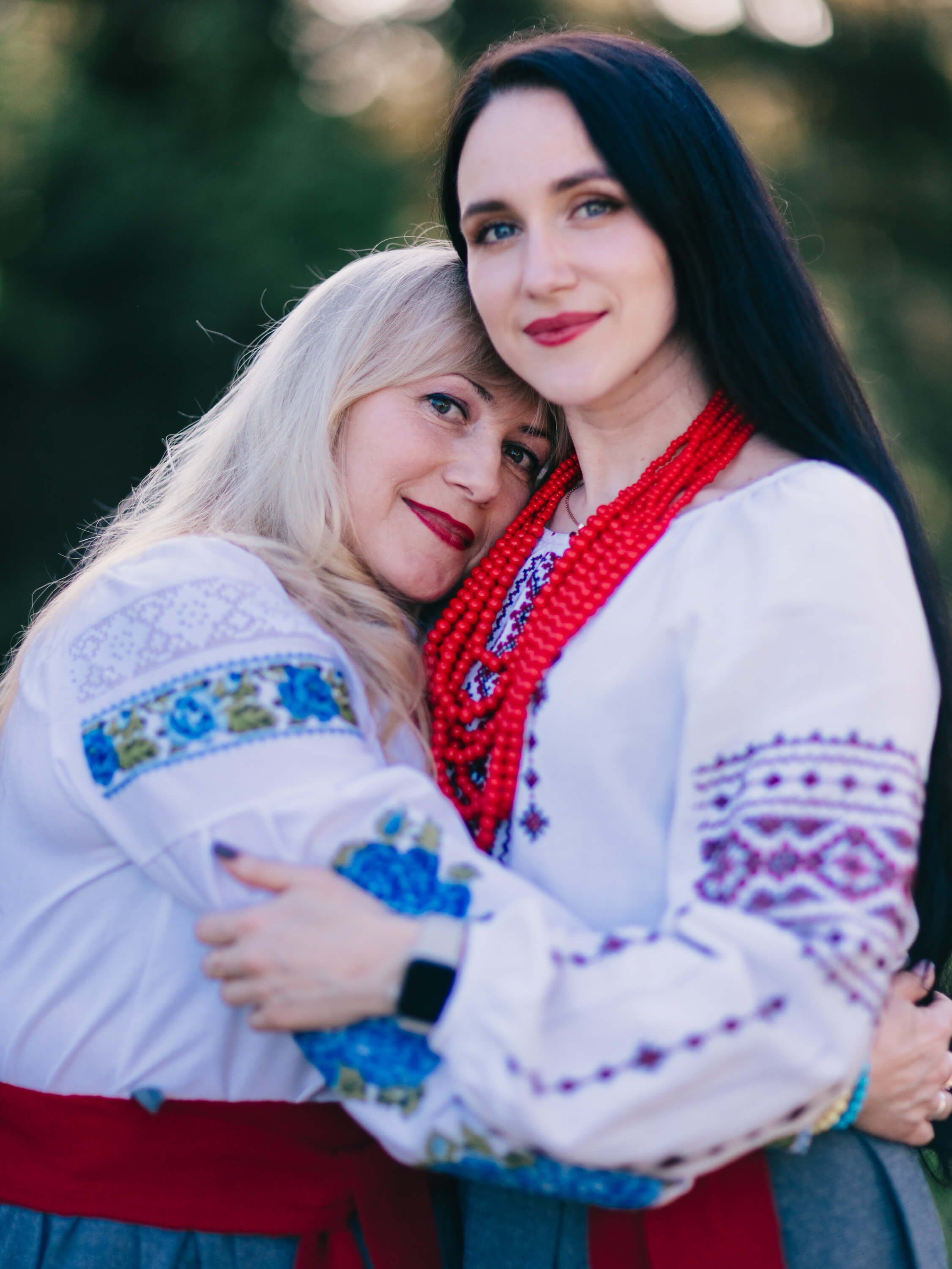 Two women embrace outdoors wearing embroidered white linen shirts, warm golden light filtering through trees behind them