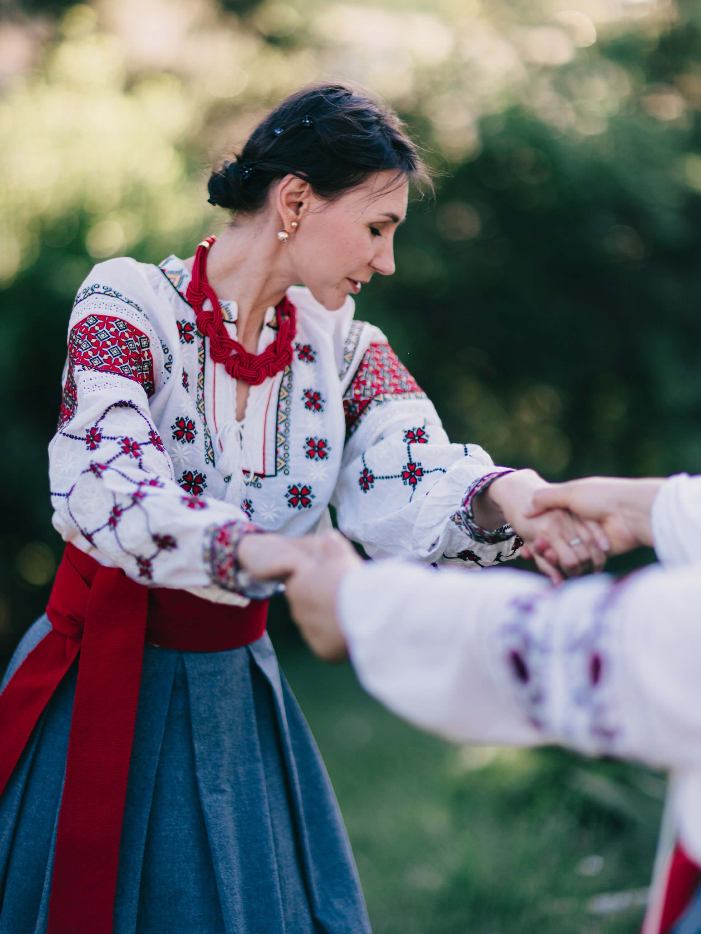 Woman in embroidered Ukrainian dress and red ribbon shakes hands with someone, golden-hour light filtering through trees behind her