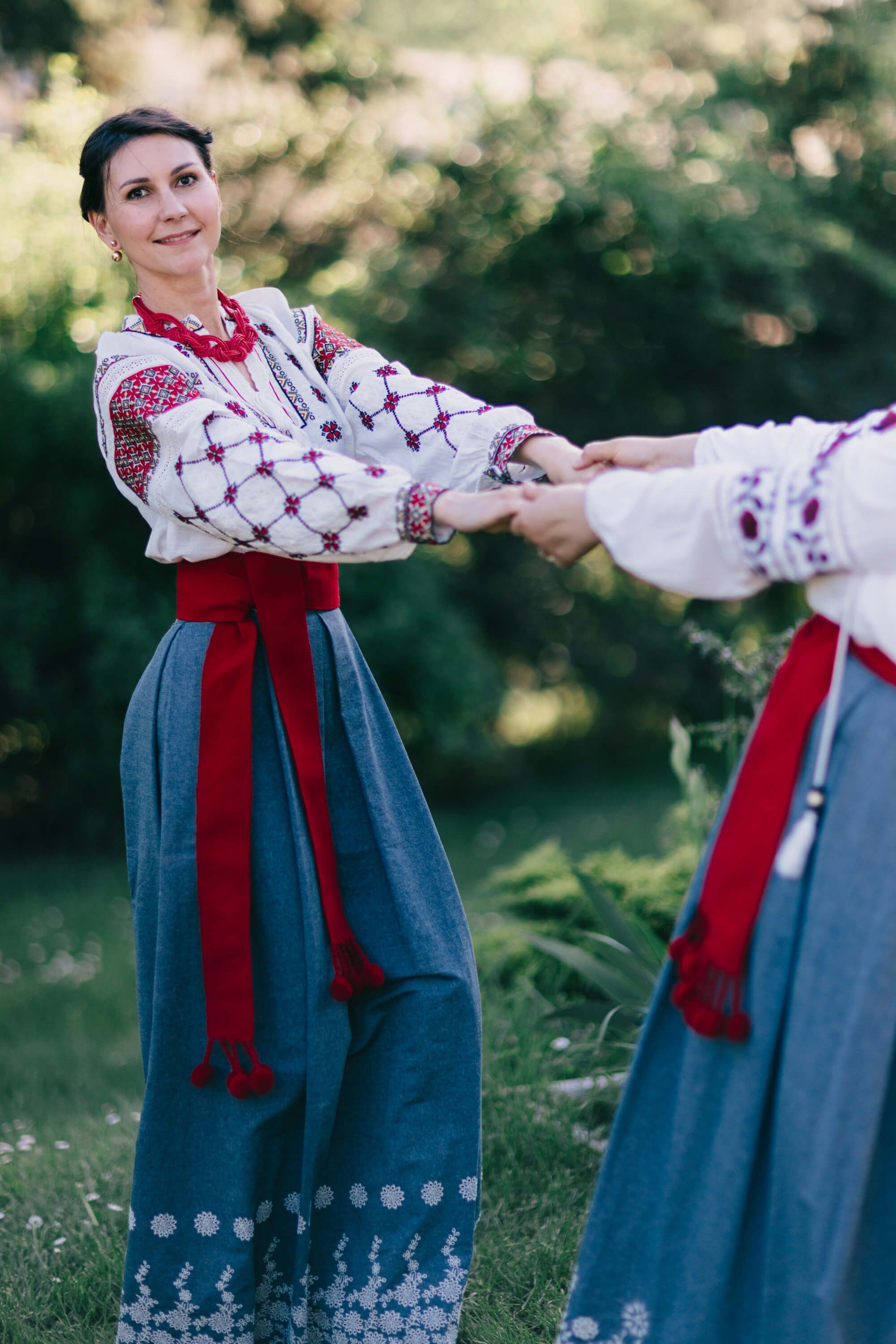 Two women in embroidered Ukrainian traditional dress hold hands in golden afternoon light, their red sashes and white lace-trimmed skirts vivid against soft…