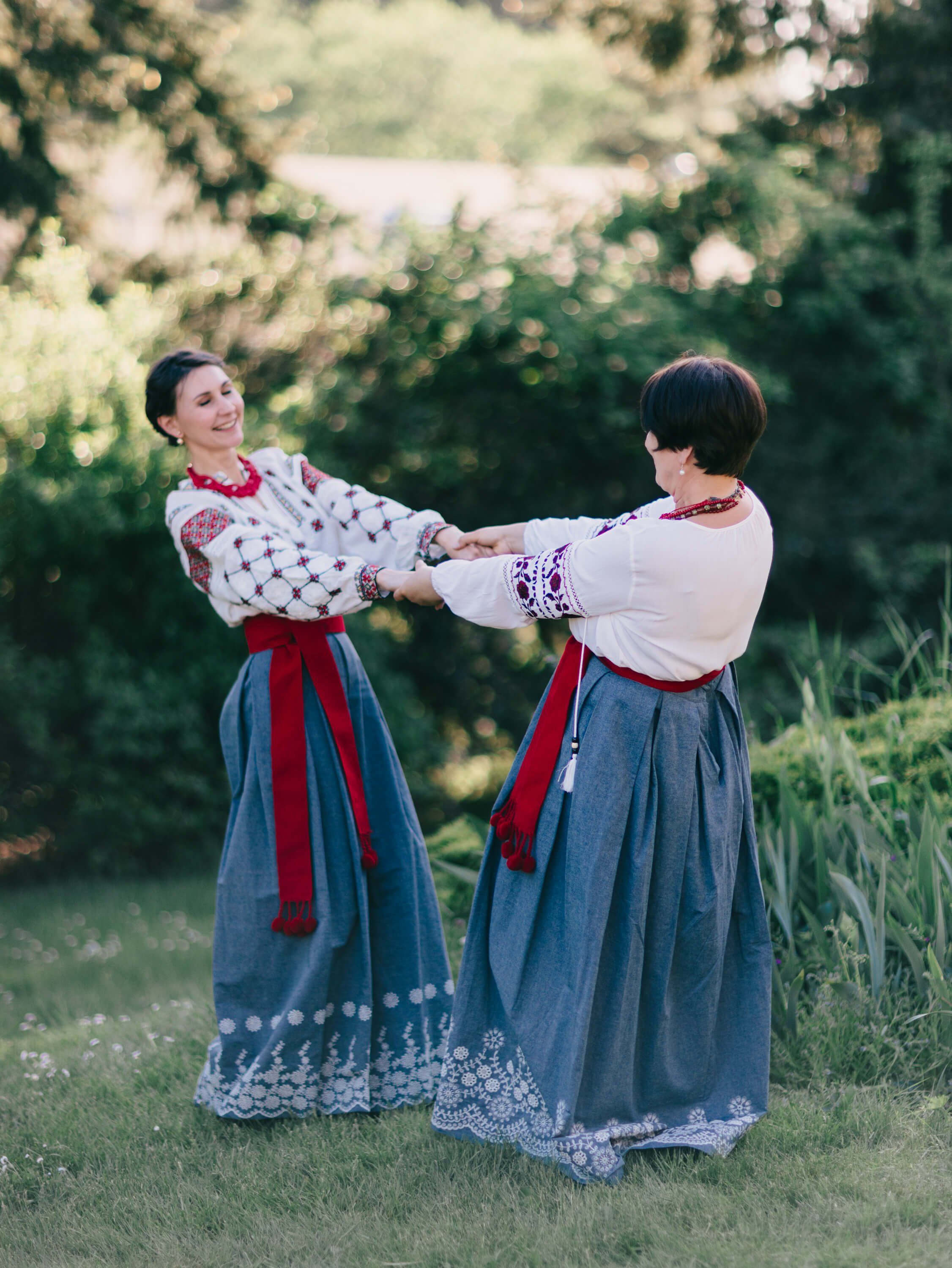 Two women in traditional Ukrainian embroidered blouses and linen skirts clasp hands in a sunlit garden setting