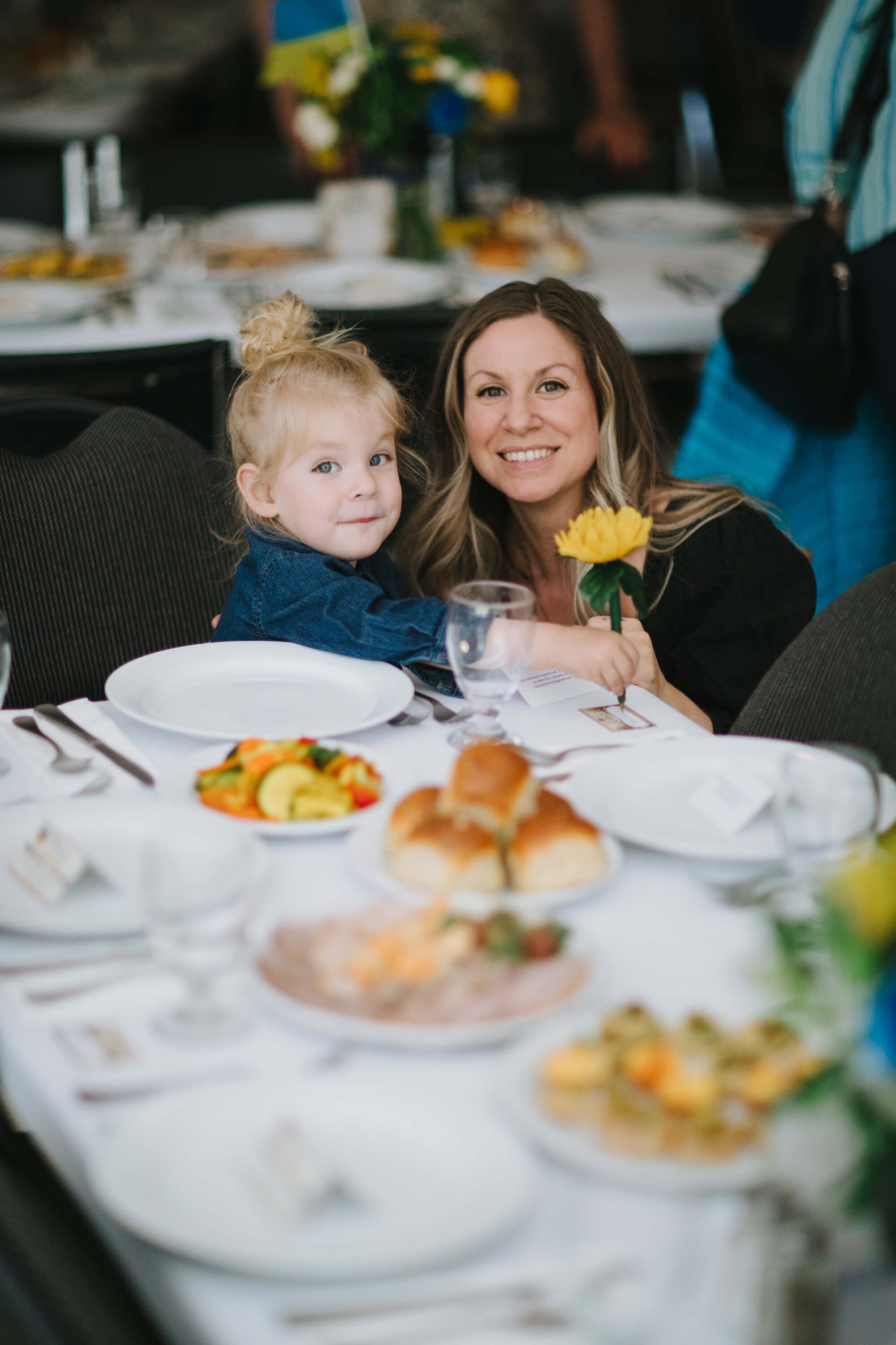 Woman and young child smile at elegant dinner table set with yellow flowers and plated food