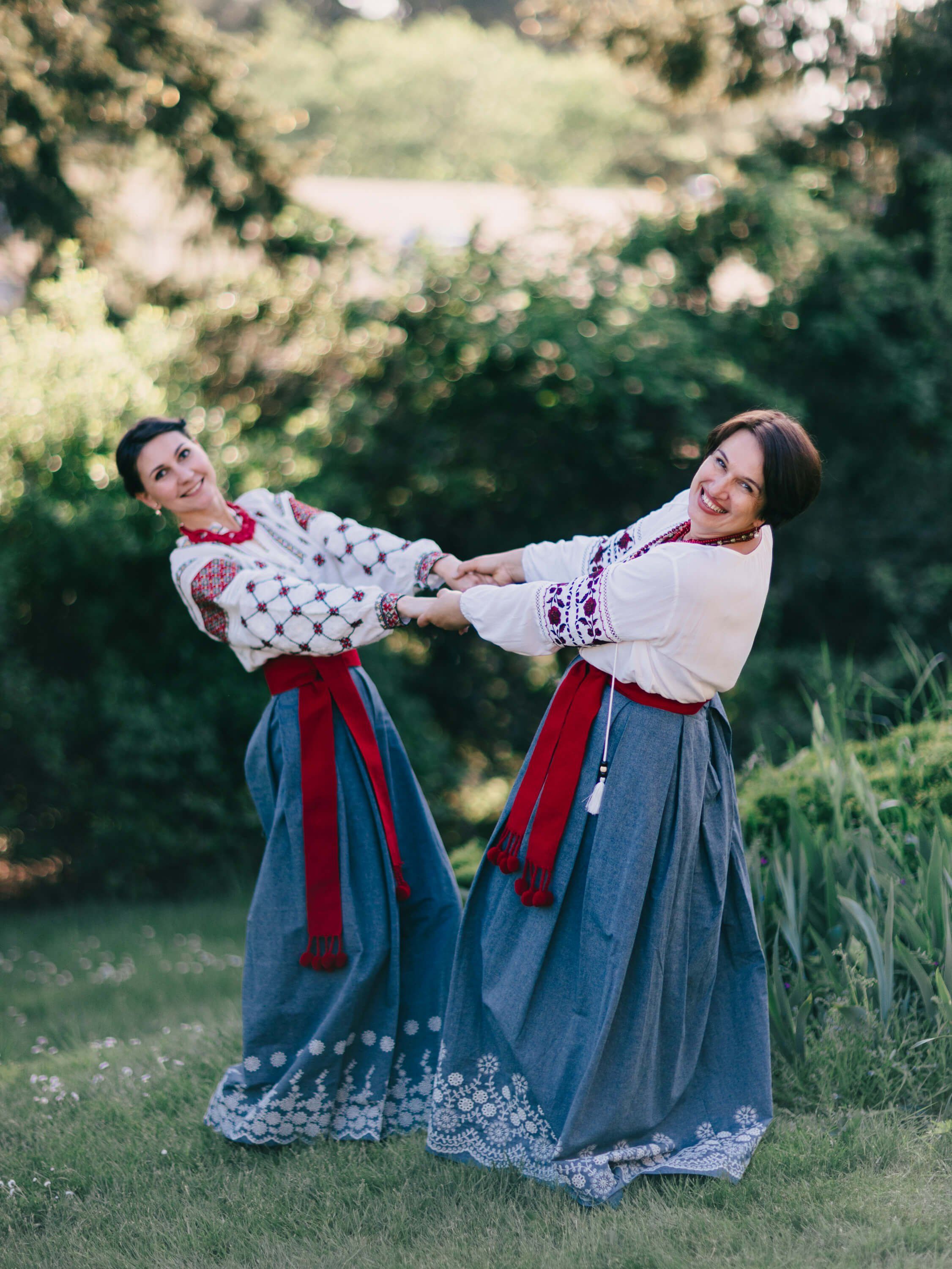 Two women in traditional Ukrainian embroidered blouses and denim skirts dance joyfully in golden evening light among green grass and trees