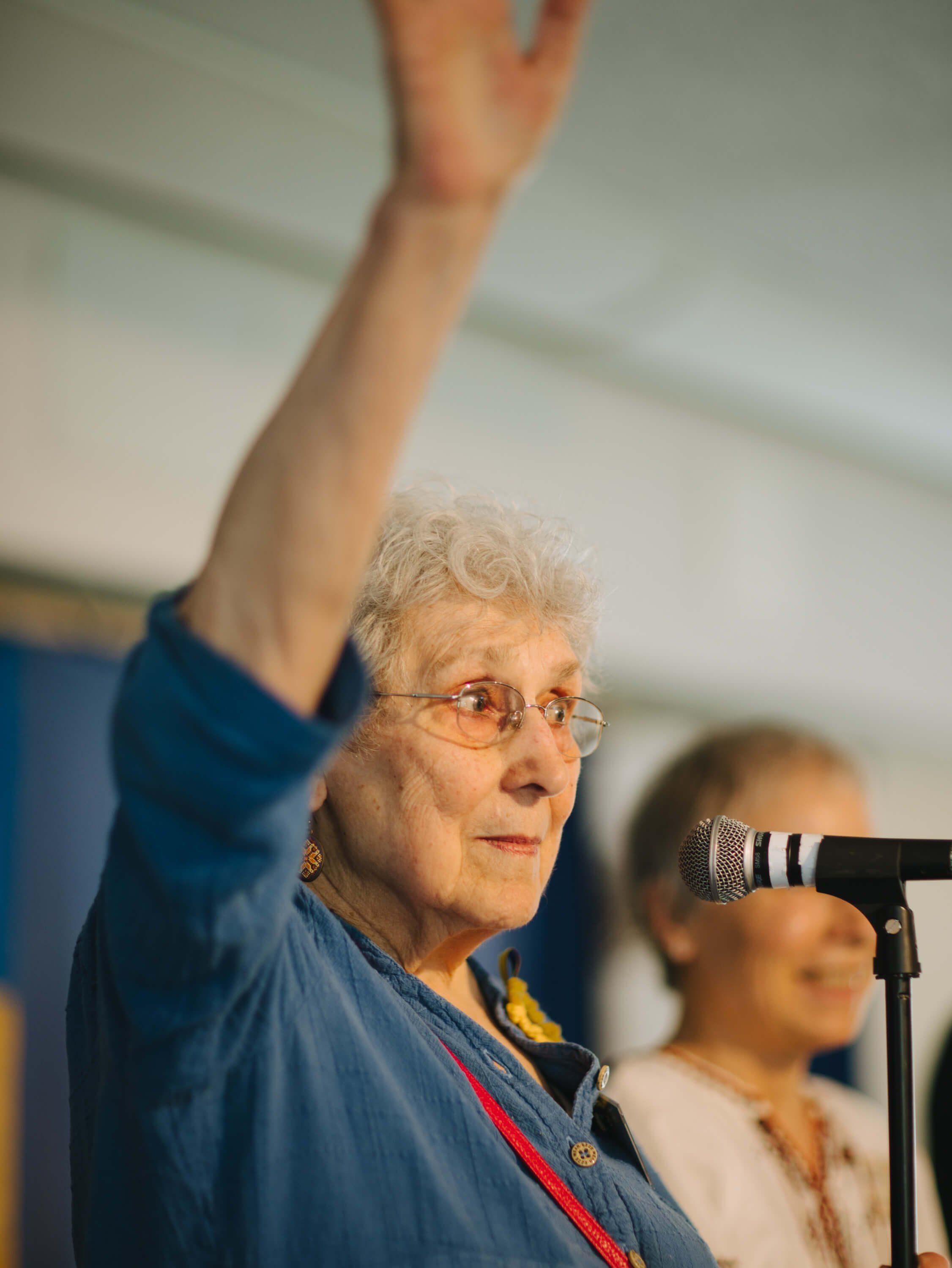 Older woman with white hair and glasses speaking passionately at a microphone, fist raised, at an indoor fundraiser event
