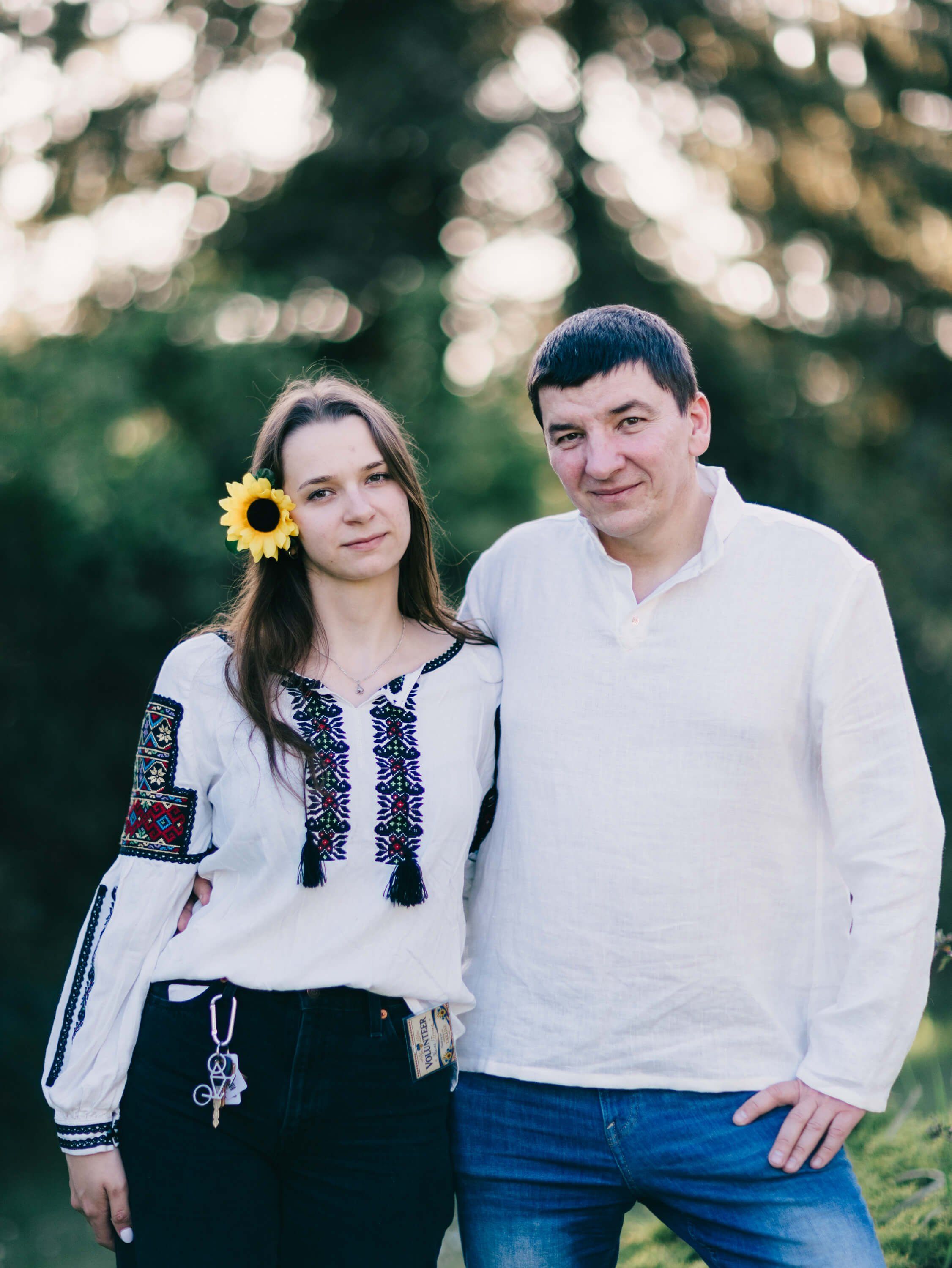 Couple in traditional Ukrainian embroidered shirt and white linen pose outdoors at golden hour, woman wearing sunflower in hair