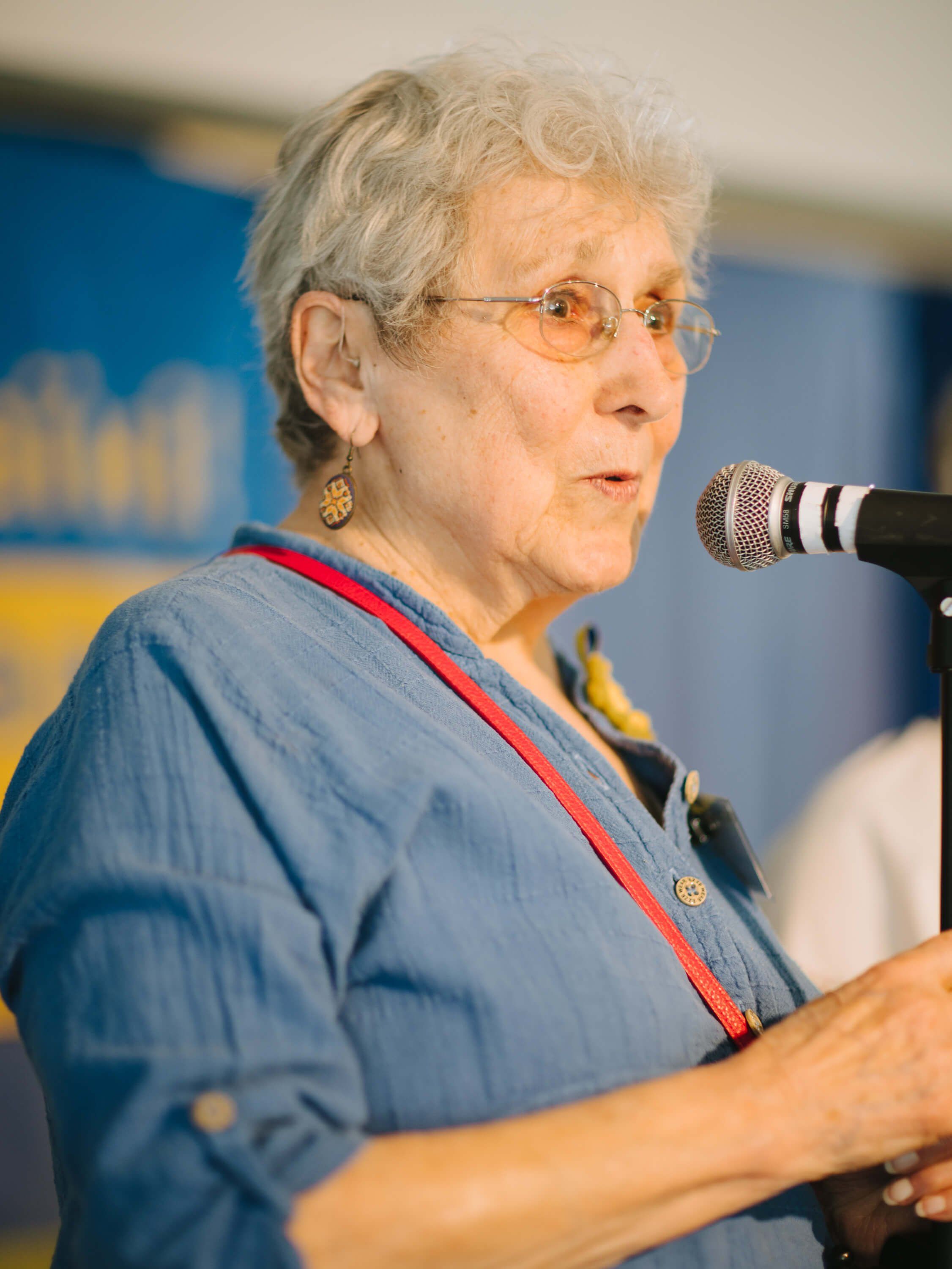 Elderly woman wearing glasses and blue shirt speaks at microphone during Ukrainian fundraiser dinner event