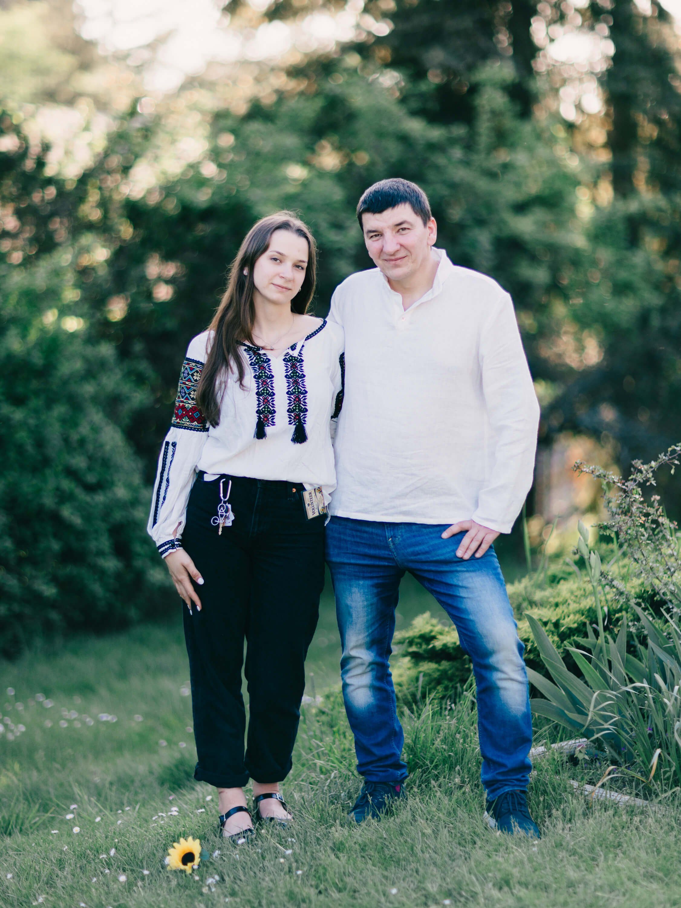 Couple in casual attire standing together on a grass field with evergreen trees and golden hour light in the background