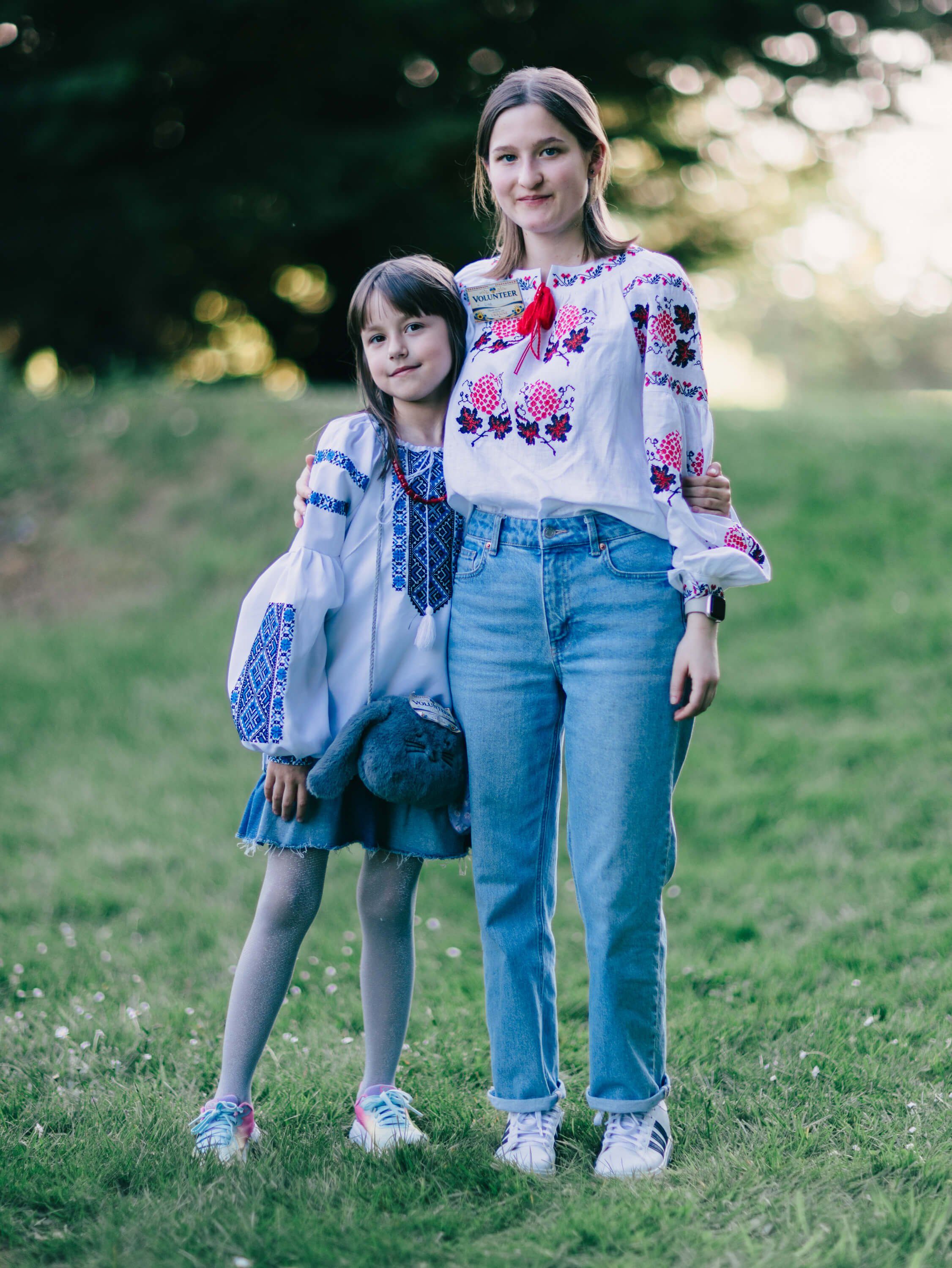 Woman and young girl wearing embroidered Ukrainian shirts stand together on grass at golden hour