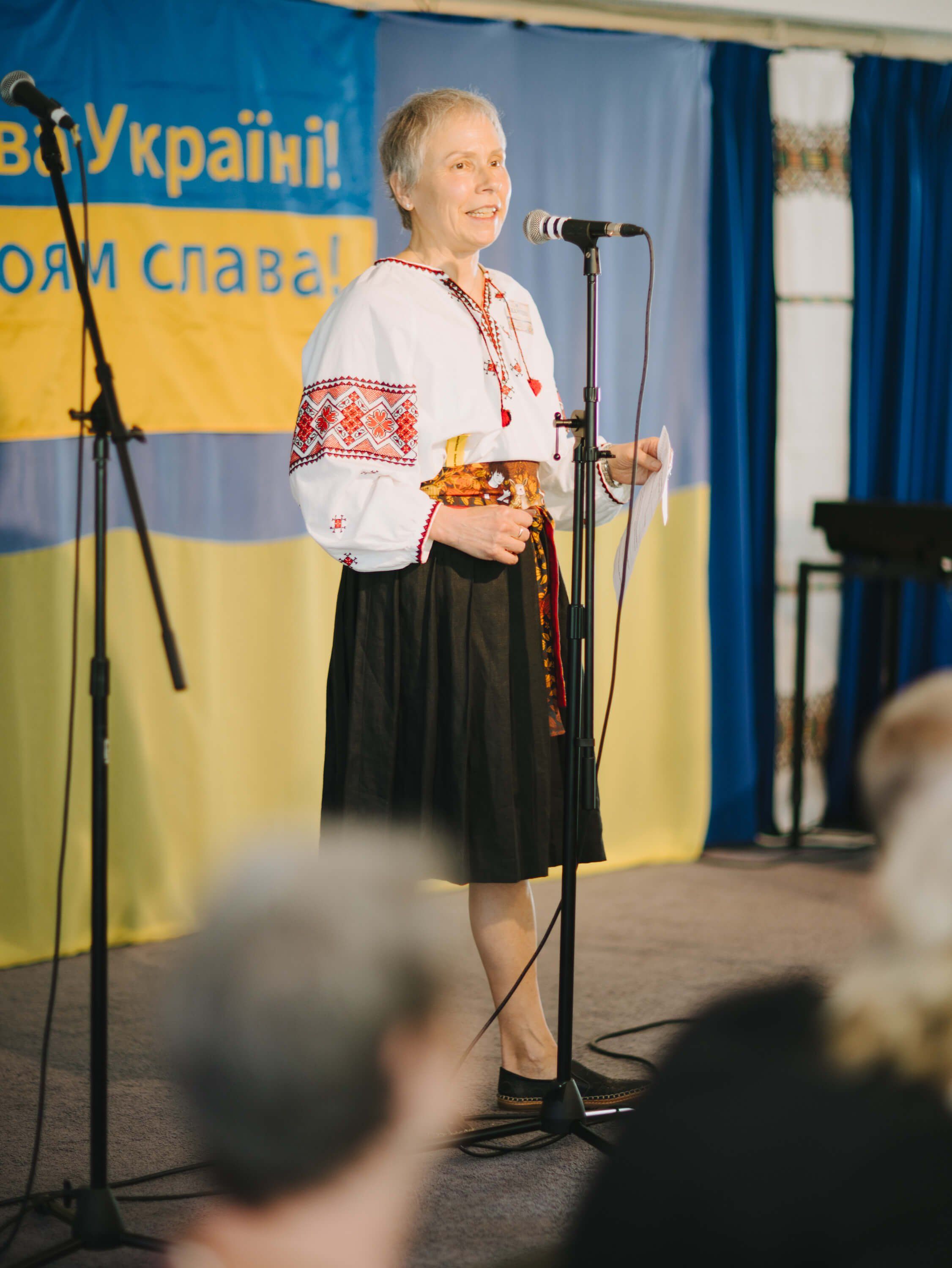 Woman in traditional Ukrainian embroidered shirt and black skirt speaks at microphone before blue and yellow Ukrainian flag backdrop
