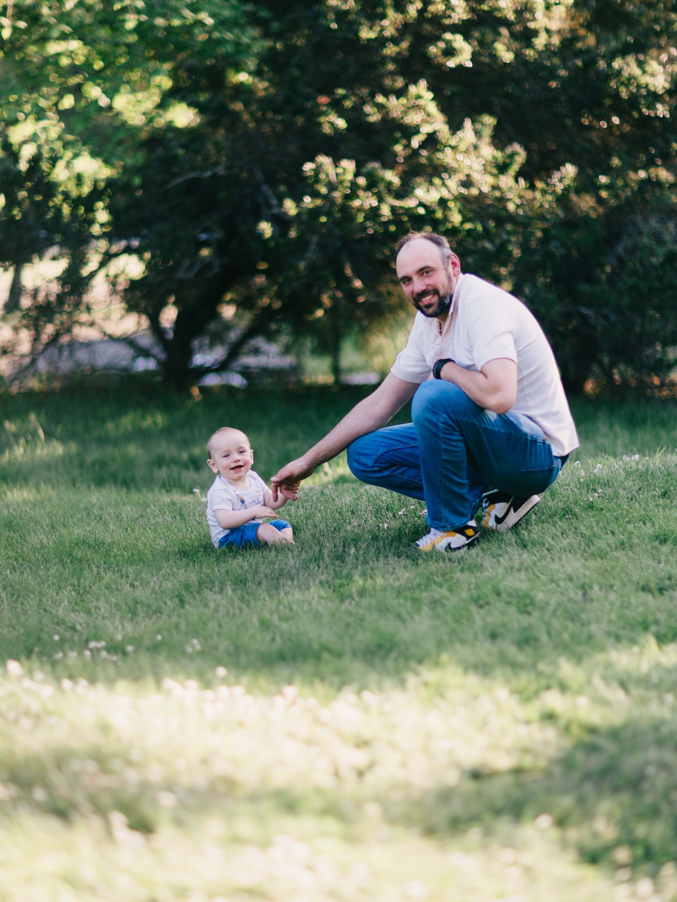 Father crouches beside his toddler on a sunlit lawn, reaching toward the child in dappled tree shade