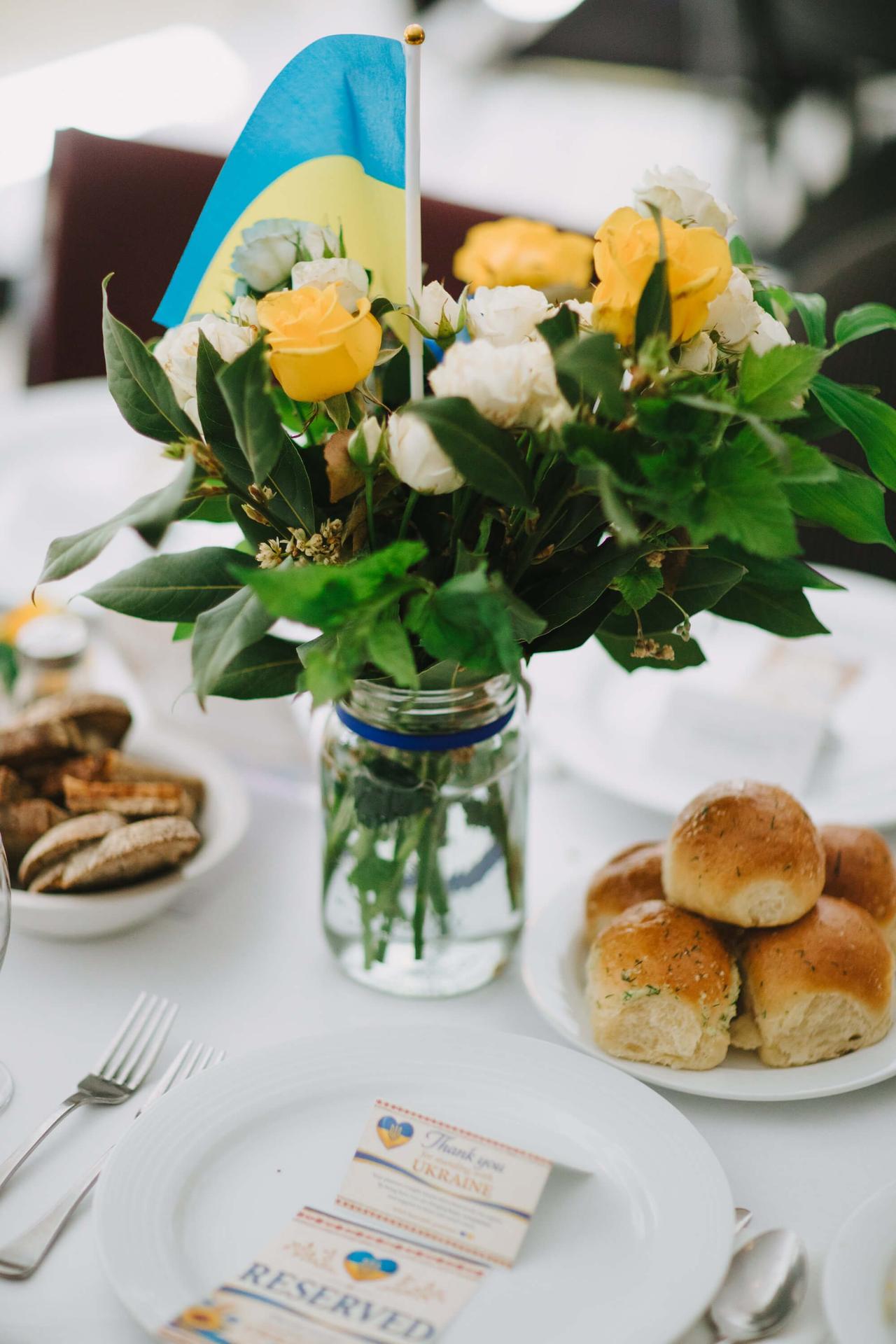 White roses in a glass vase with blue and yellow ribbon stand beside Ukrainian fundraiser dinner cards on a white surface