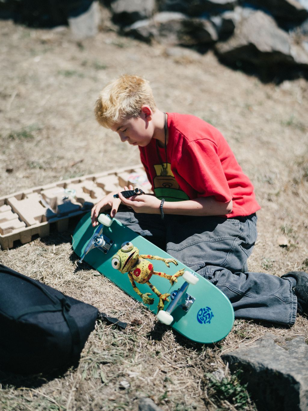 Young skateboarder in red shirt examines grip tape on turquoise skateboard at Kanaka Road Skatepark