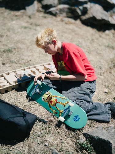 Young skateboarder in red shirt examines grip tape on turquoise skateboard at Kanaka Road Skatepark