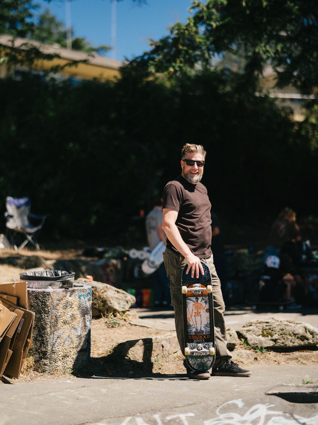 Bearded man in sunglasses holds skateboard at Kanaka Road Skatepark with evergreen hedge backdrop