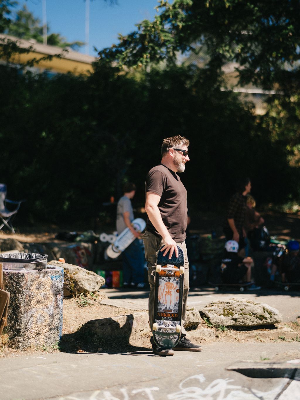Man in brown shirt stands with skateboard at concrete skate park on sunny day, trees and spectators visible in background