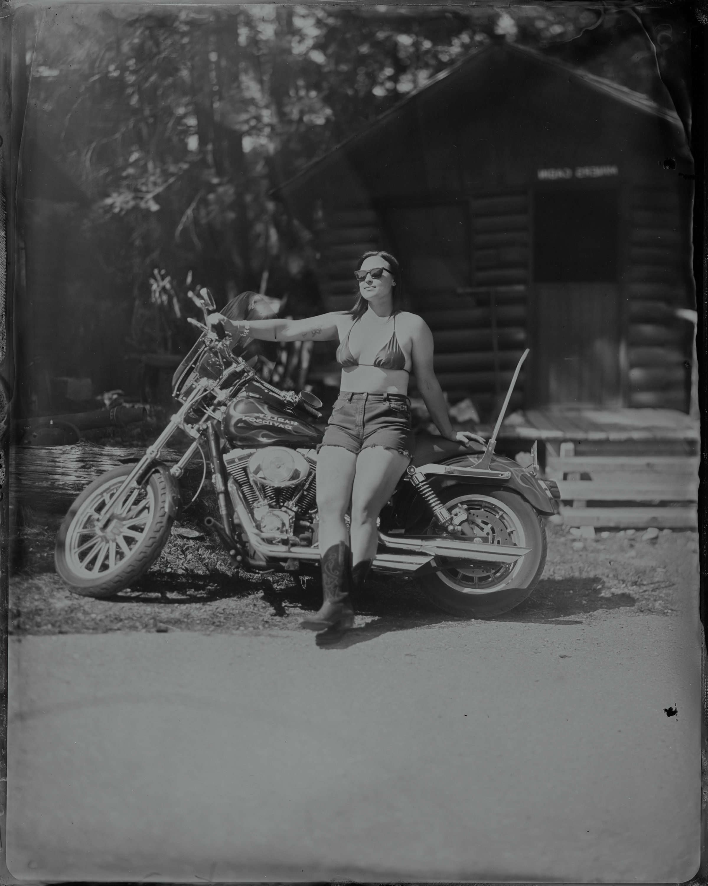 Woman in bikini top and shorts posing against classic motorcycle, wooden cabin and trees visible behind in black and white