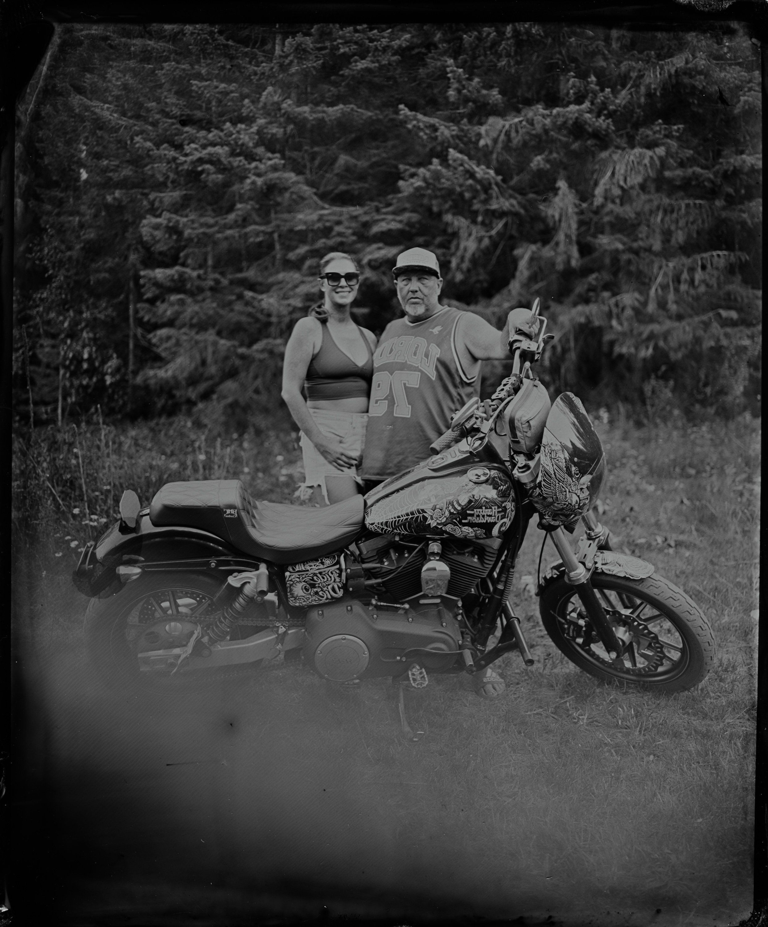 Couple posing with a motorcycle and sidecar against evergreen trees, black and white photograph with soft natural lighting