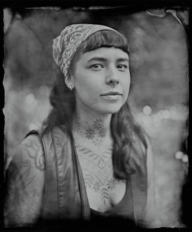 Close profile of a person wearing a patterned headwrap, with decorative tattoos visible on neck and chest against dark background