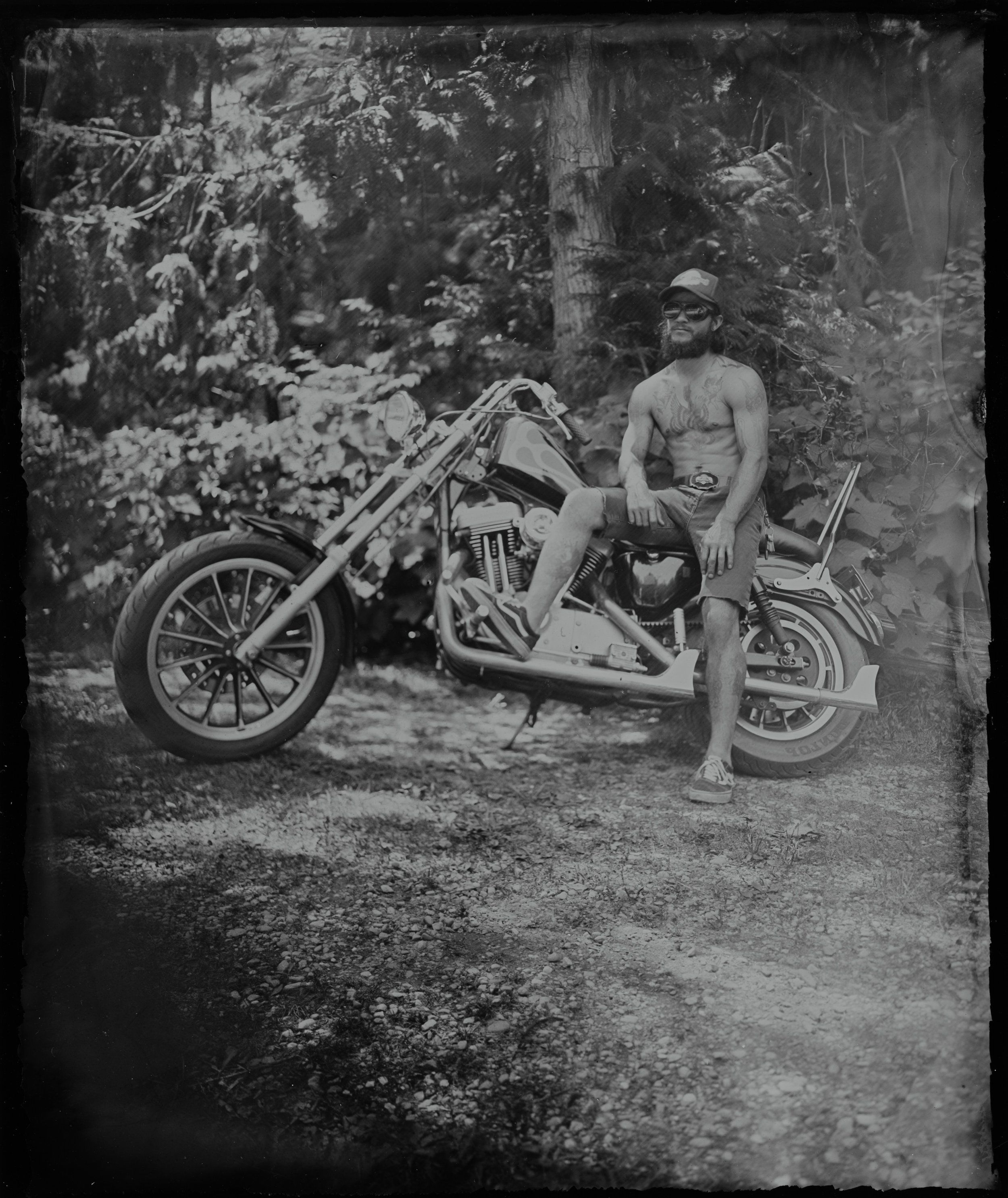 Shirtless man on custom chopper motorcycle in forest clearing, black and white vintage photograph