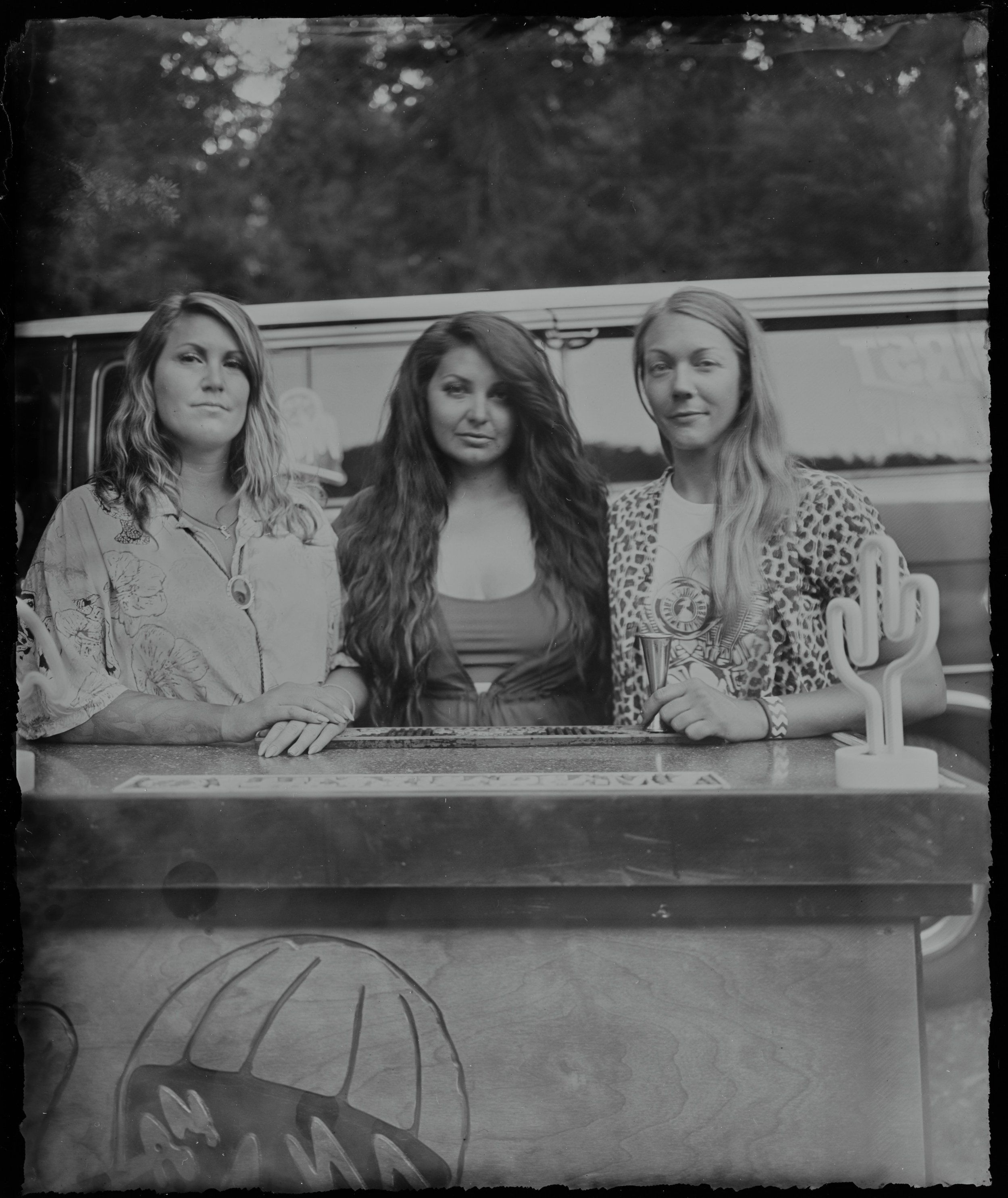Three women lean against the side of a vehicle, dressed in vintage 1970s clothing, gazing thoughtfully off-camera in this black and white portrait