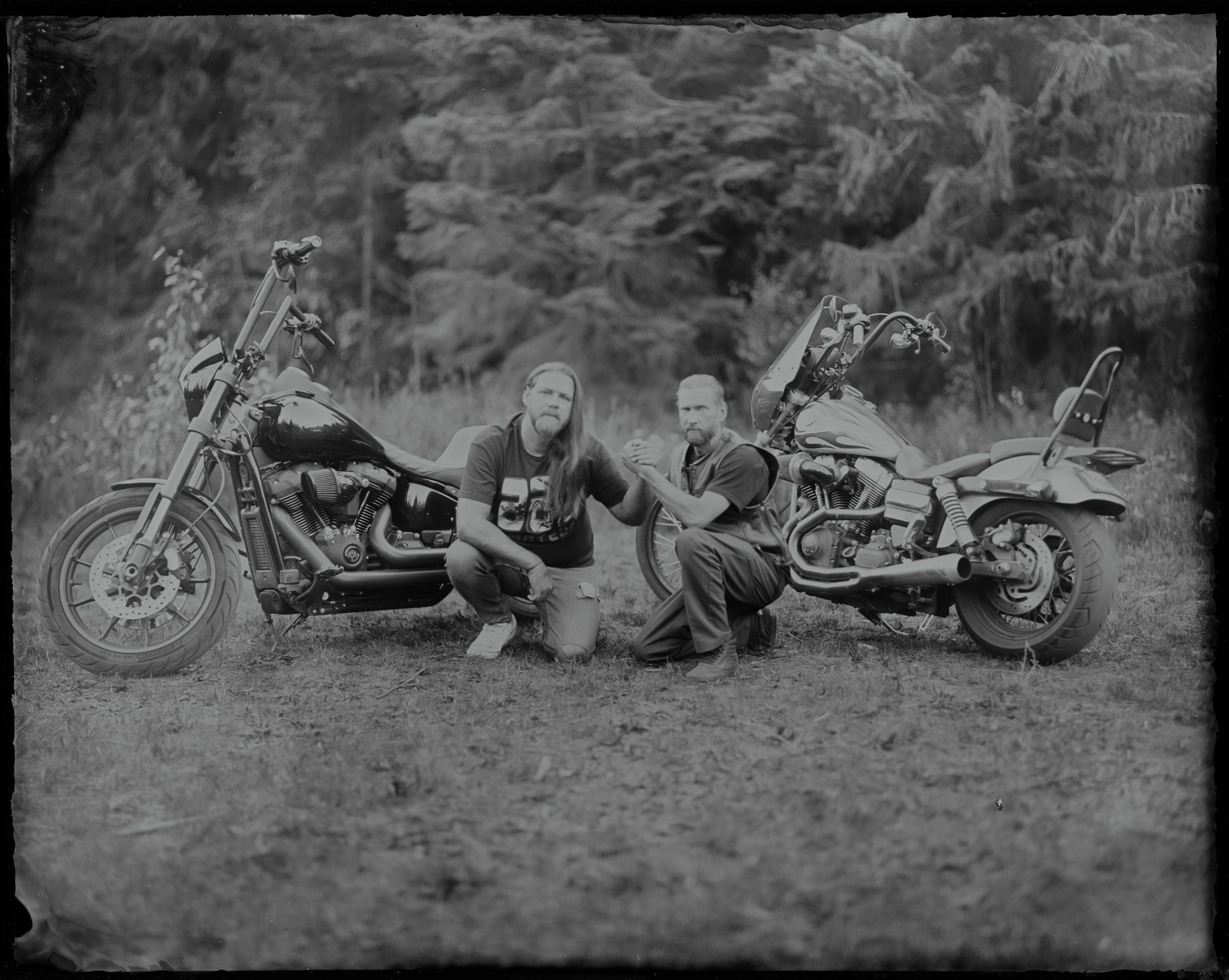 Two leather-clad bikers crouch beside their motorcycles on a dusty road, surrounded by forest, in moody black and white