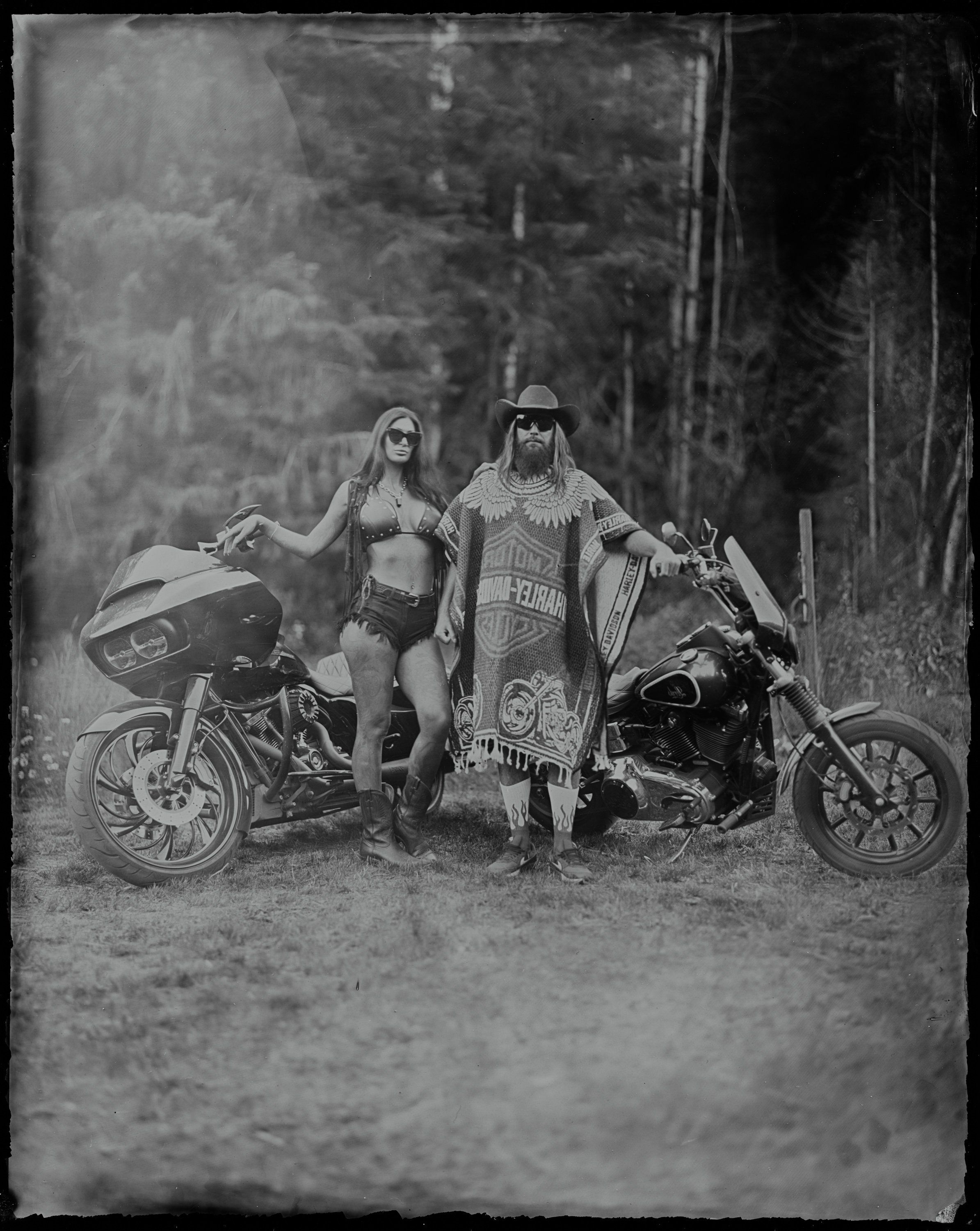 Two women in bohemian style clothing pose between two motorcycles on a forest road, rendered in black and white