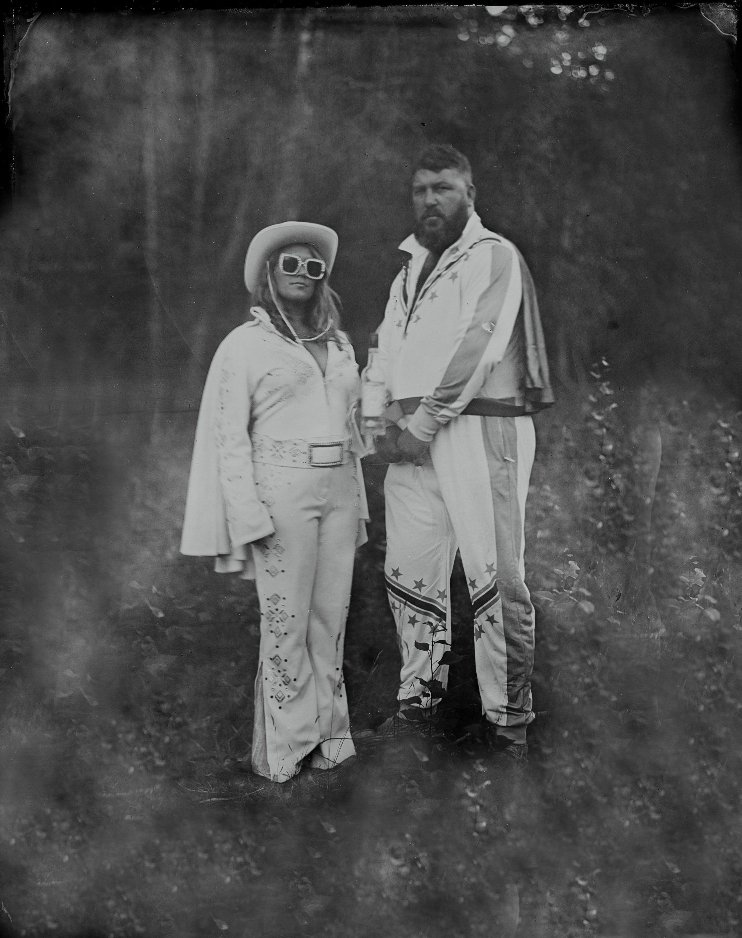 Couple in vintage-inspired white clothing standing outdoors, woman wearing large sunglasses and wide-brimmed hat, soft natural light filtering through trees