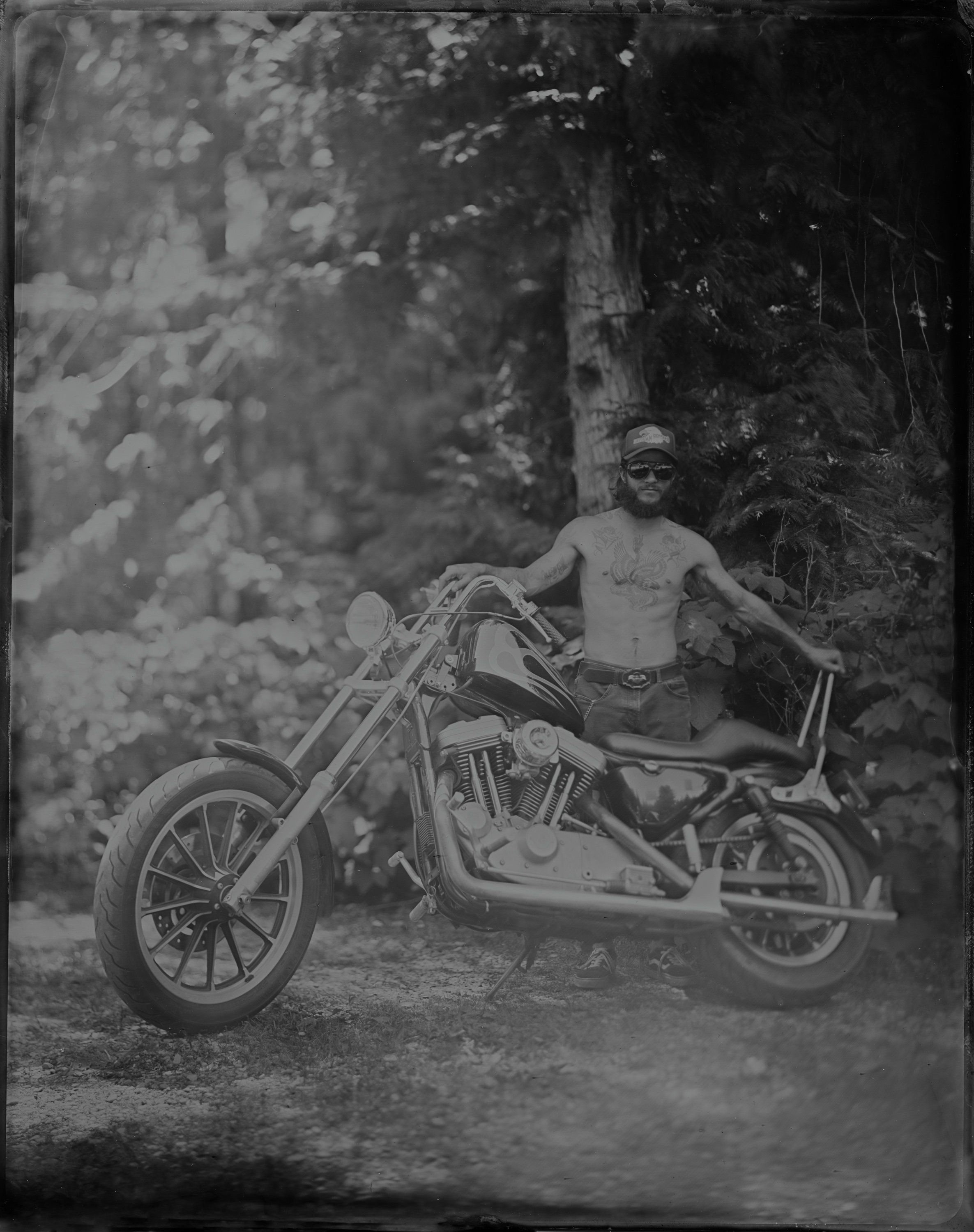 Shirtless man in hat posing with vintage motorcycle on tree-lined dirt road, black and white photograph