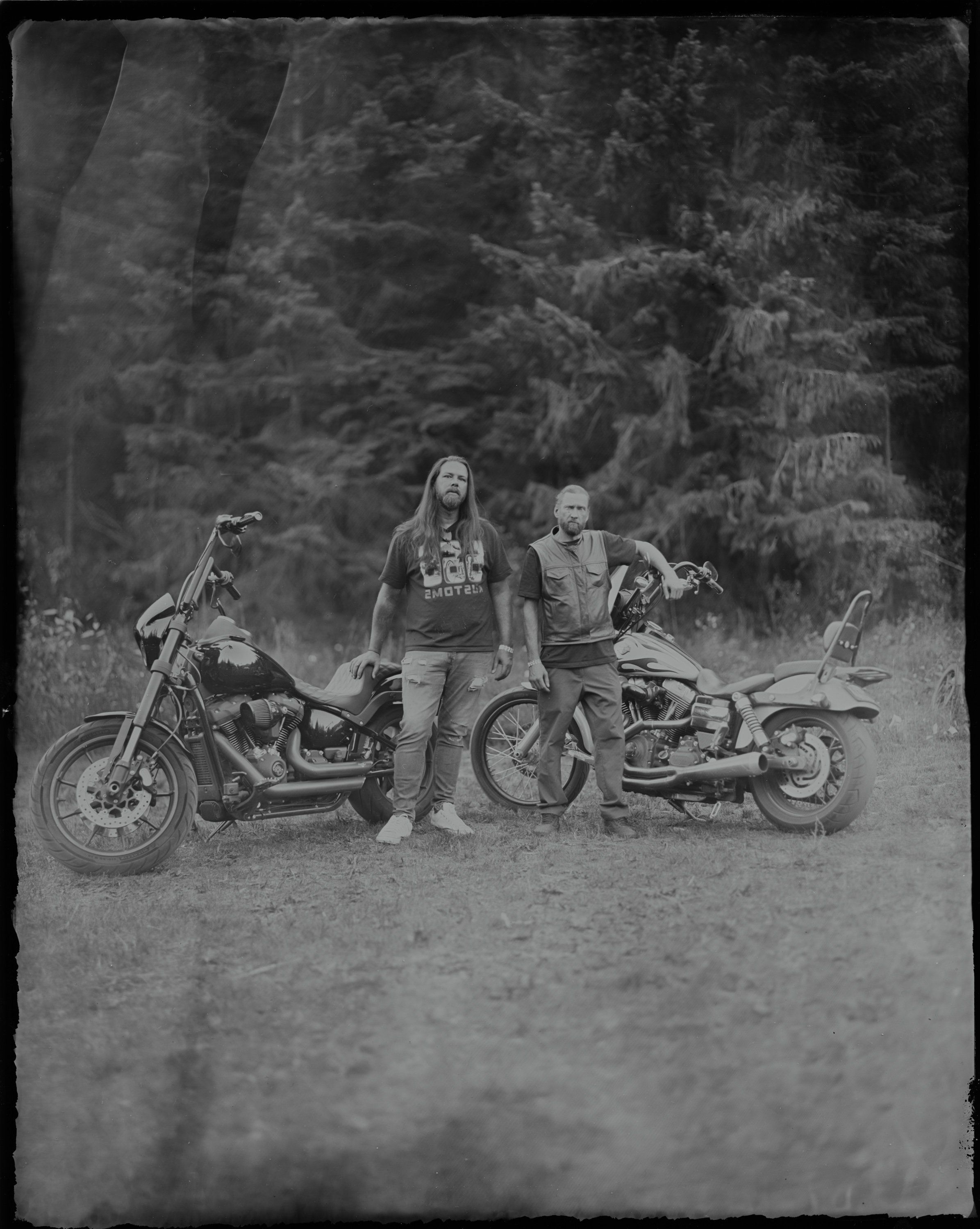 Two people stand between custom motorcycles on a forested dirt road, rendered in dramatic black and white