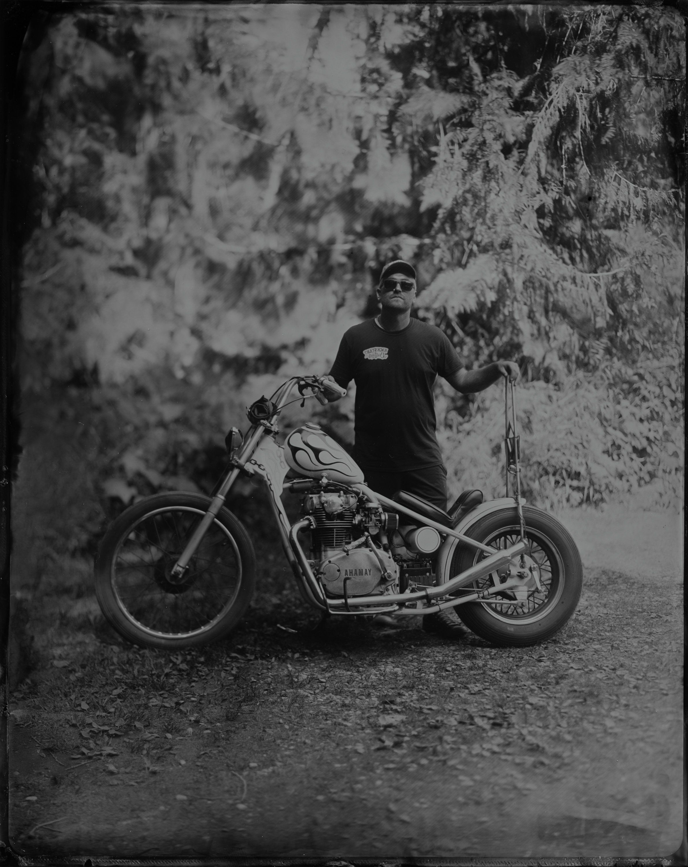Man in sunglasses stands beside a custom chopper motorcycle on a tree-lined forest road, black and white