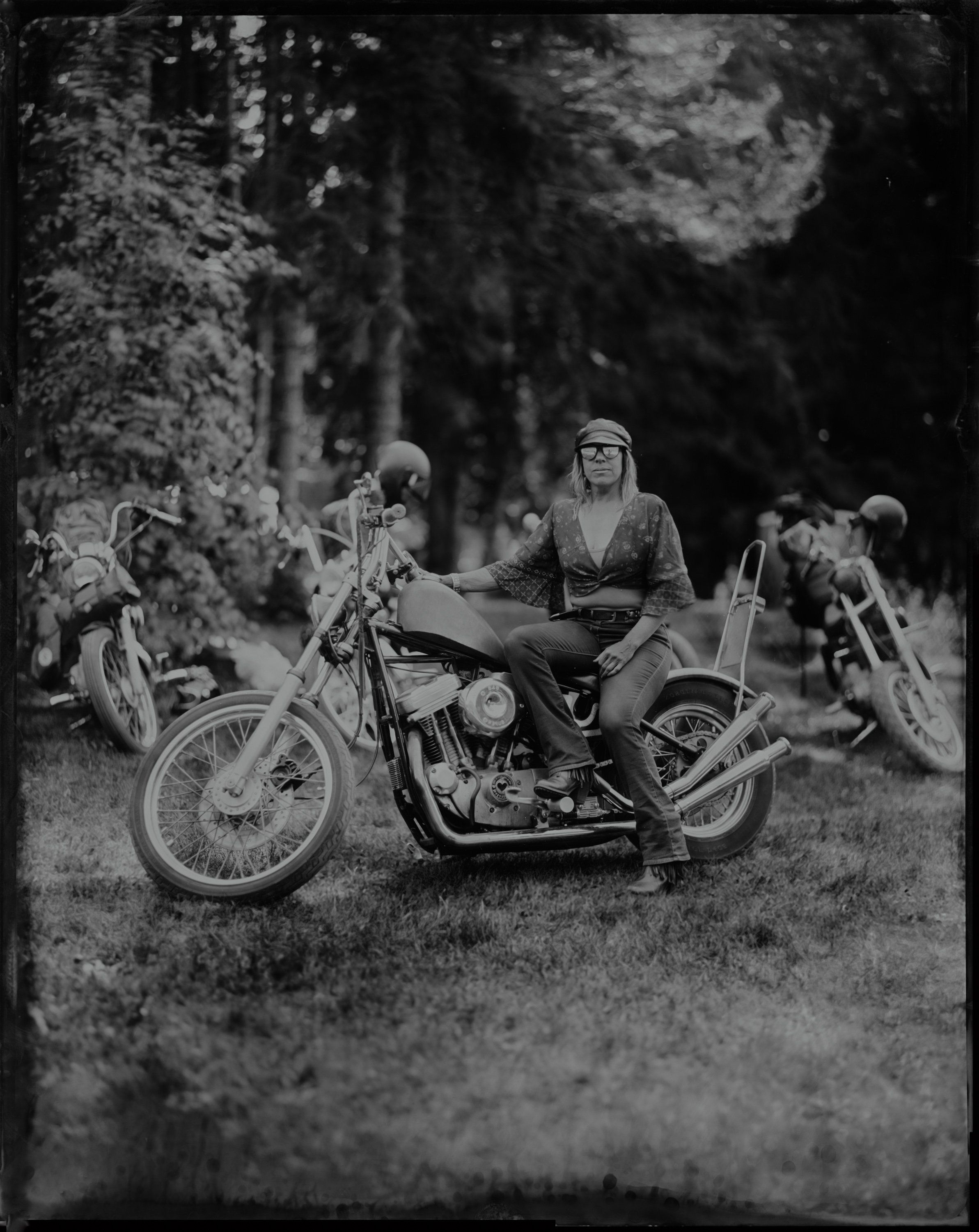Woman in vintage dress and eyeglasses seated on chopper motorcycle surrounded by leather-clad riders in dappled forest light