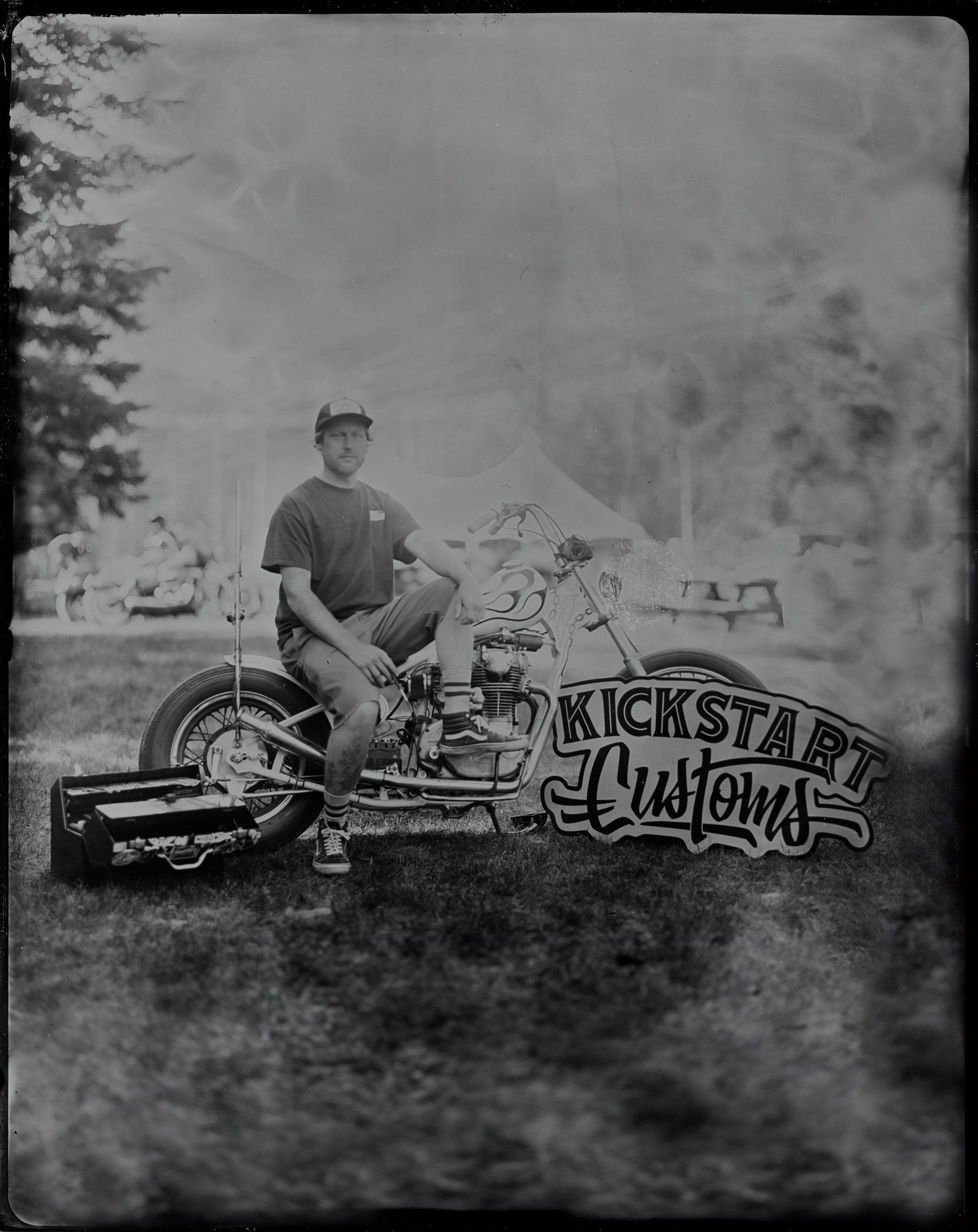 Man seated on custom motorcycle with "Kickstart Customs" signage, evergreen trees and overcast sky behind him