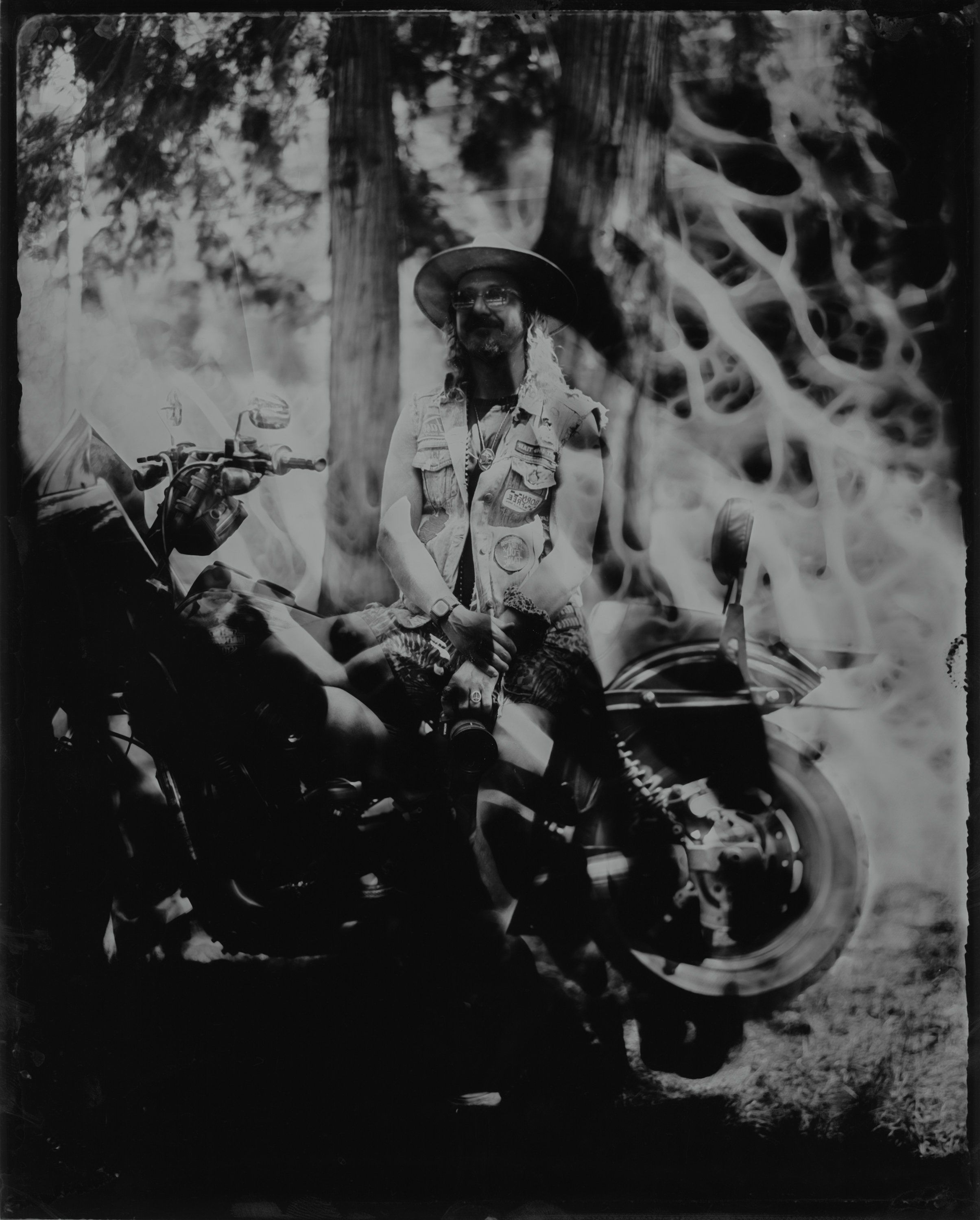 Man in cowboy hat seated on motorcycle against textured wall, dramatic shadows from foliage overhead