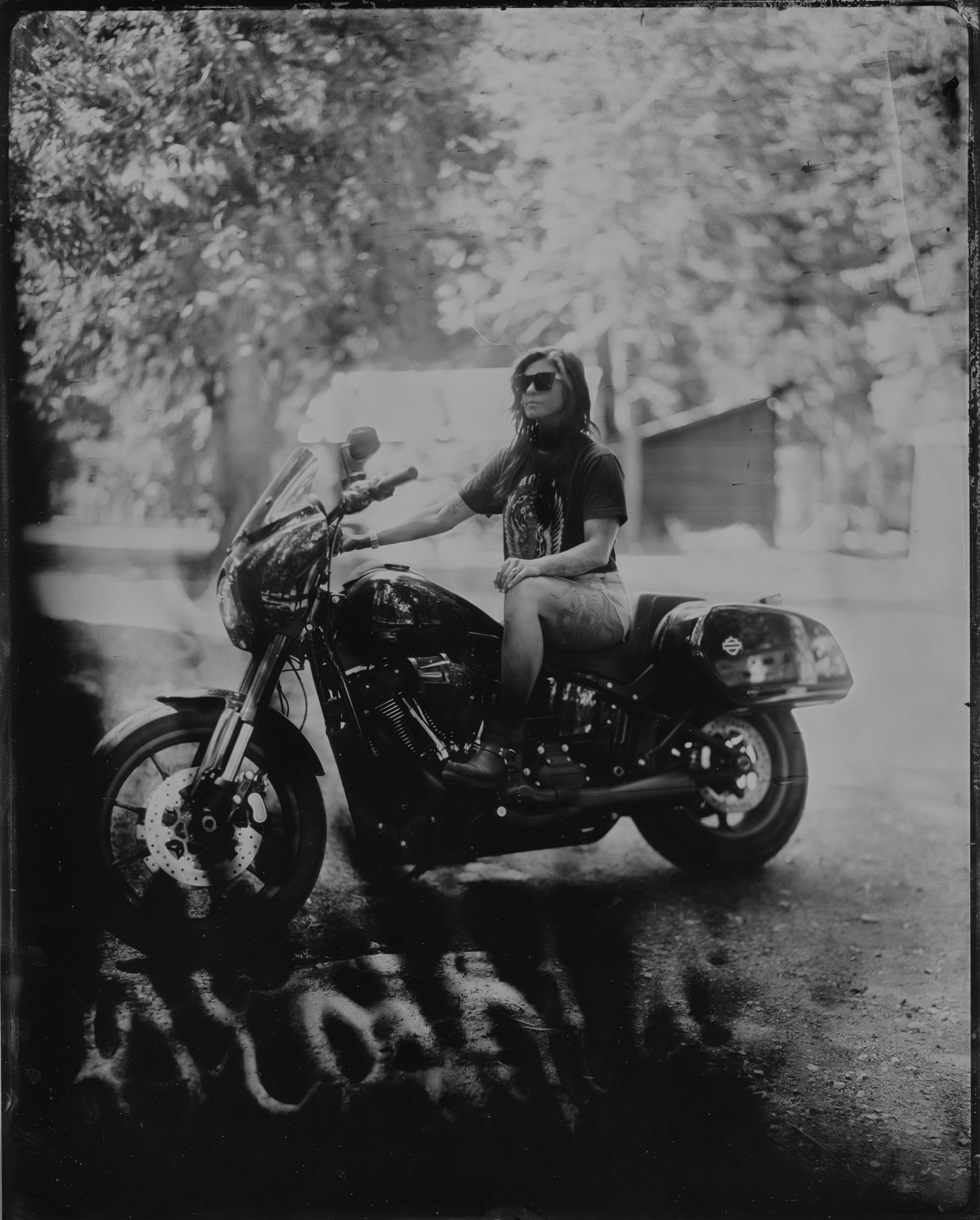 Person on motorcycle in dappled forest light, long hair flowing, hand raised in relaxed pose, black and white film photography