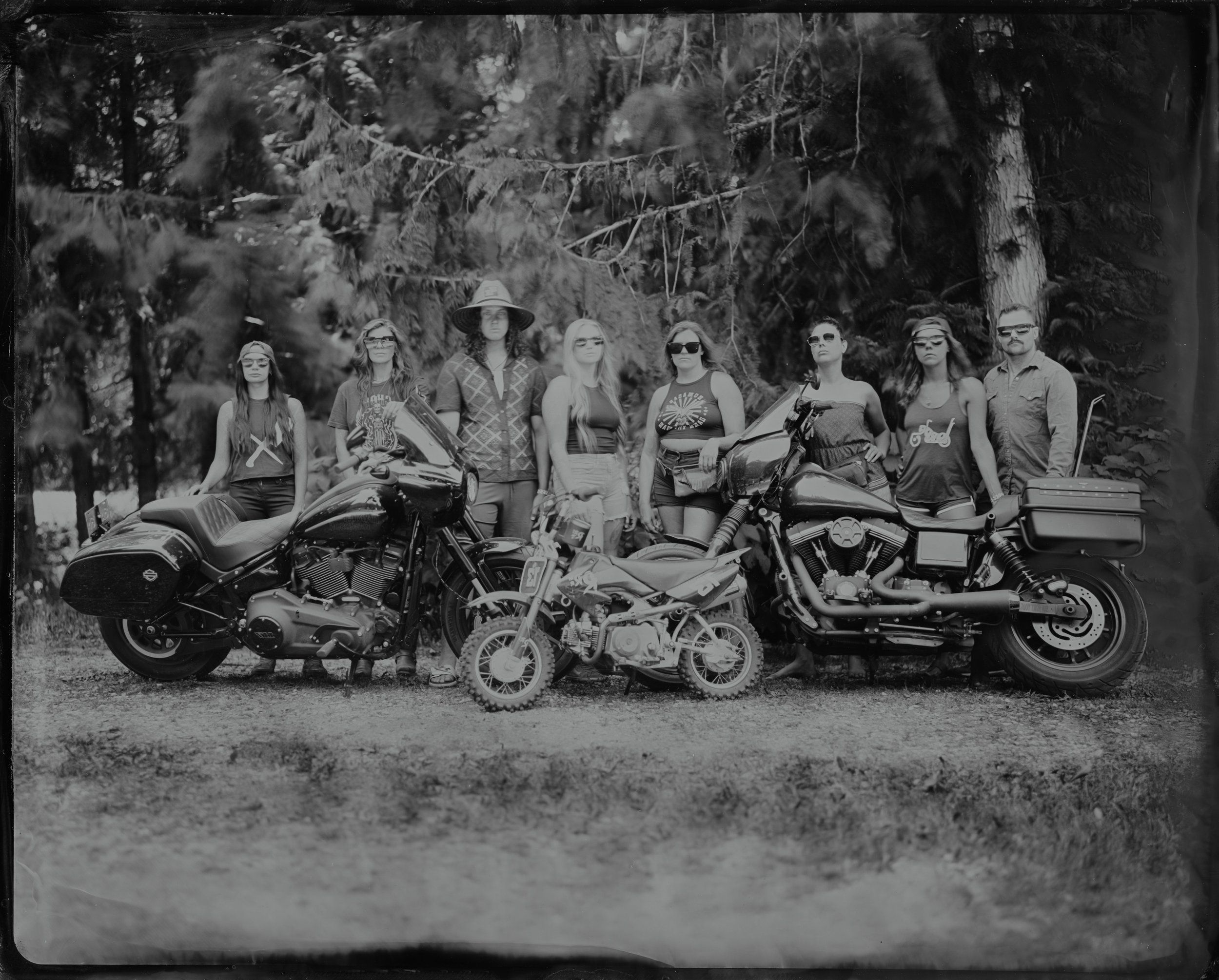 Group of leather-clad riders posed with three motorcycles among tall trees, black and white photograph