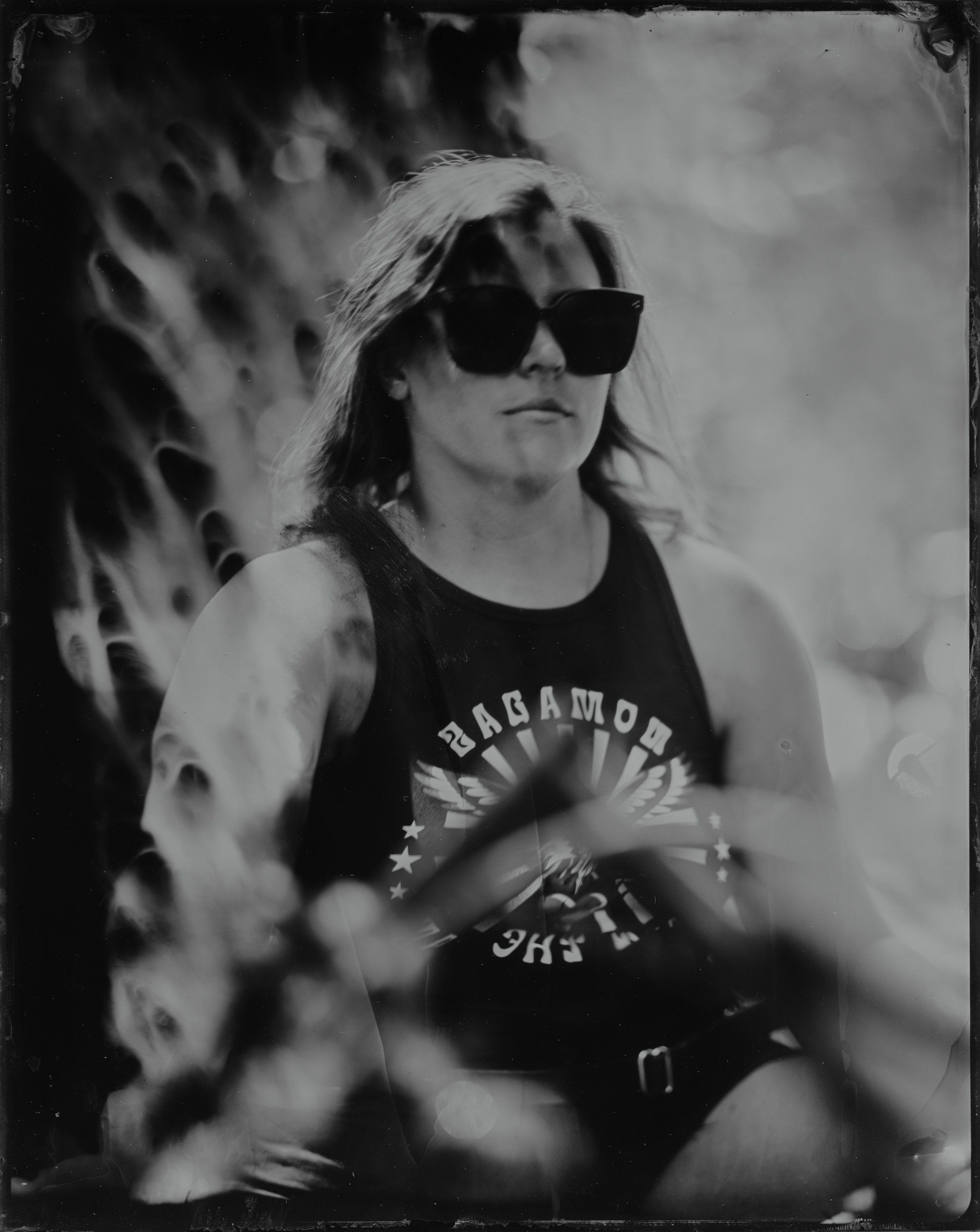 Person in dark sunglasses and band tee against blurred natural background, black and white portrait with dramatic shadows and movement