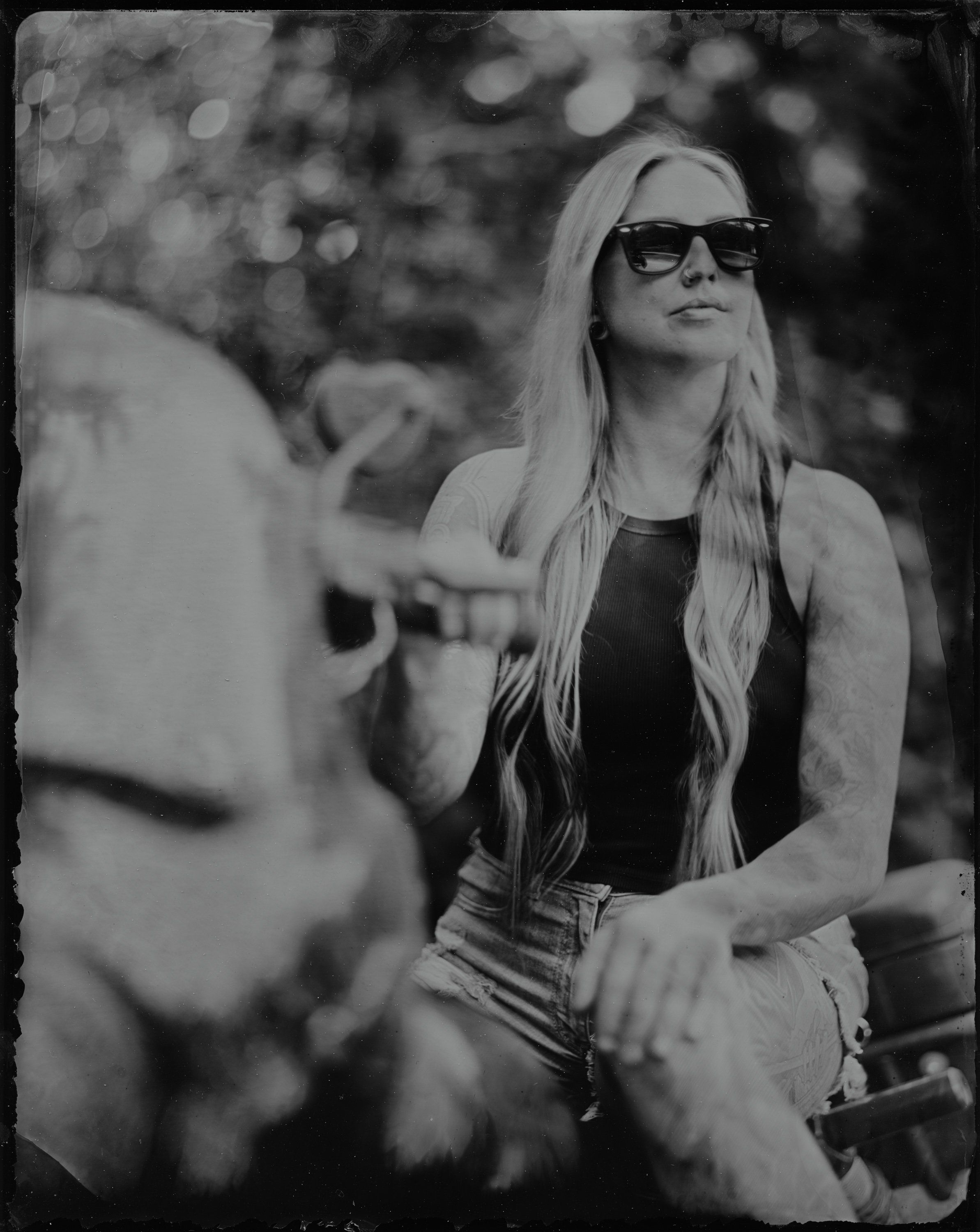Woman in sunglasses and long blonde hair gazes upward in black and white, soft bokeh foliage behind her