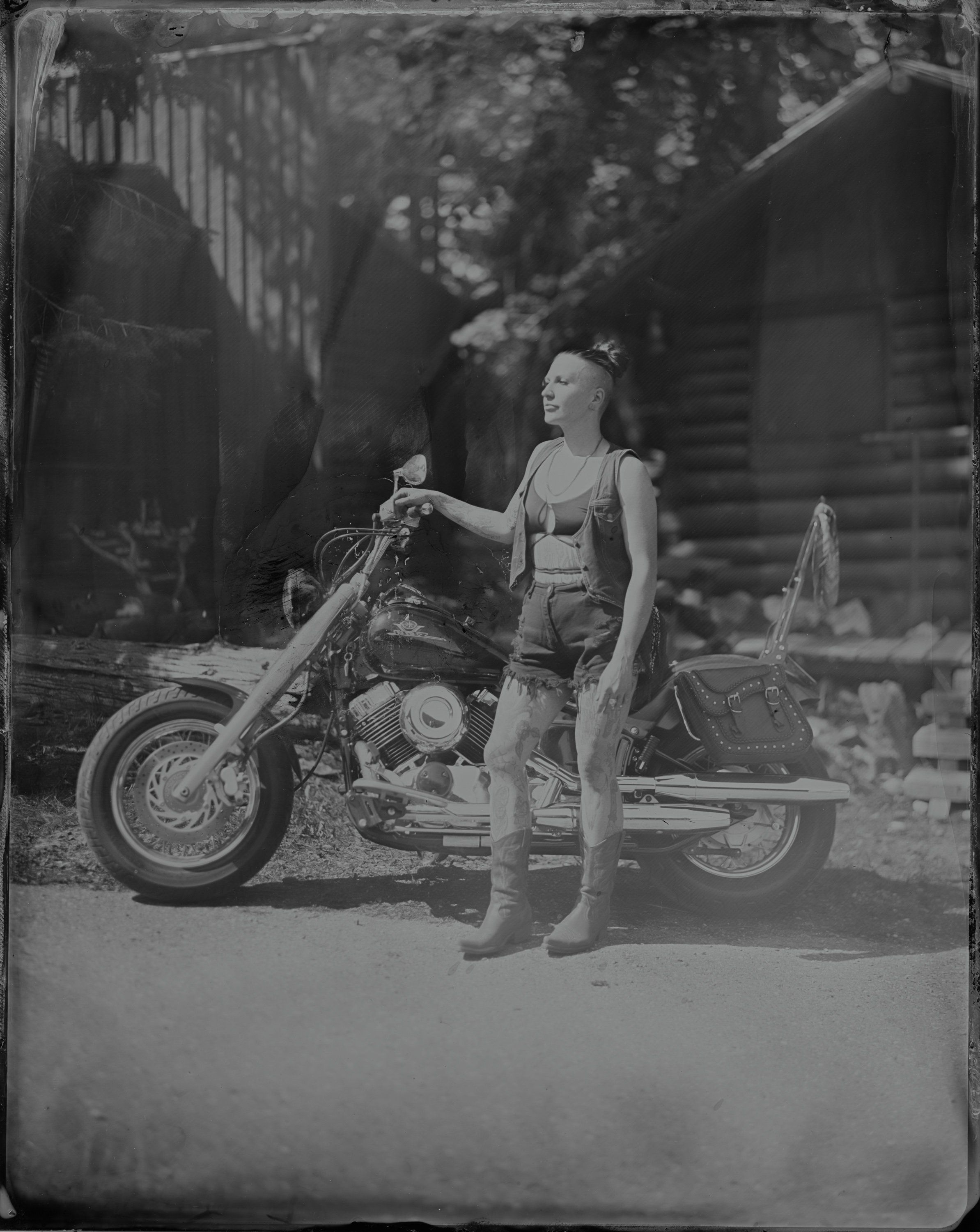 Woman in vintage swimwear stands beside a classic motorcycle, black and white portrait with natural woodland setting behind her