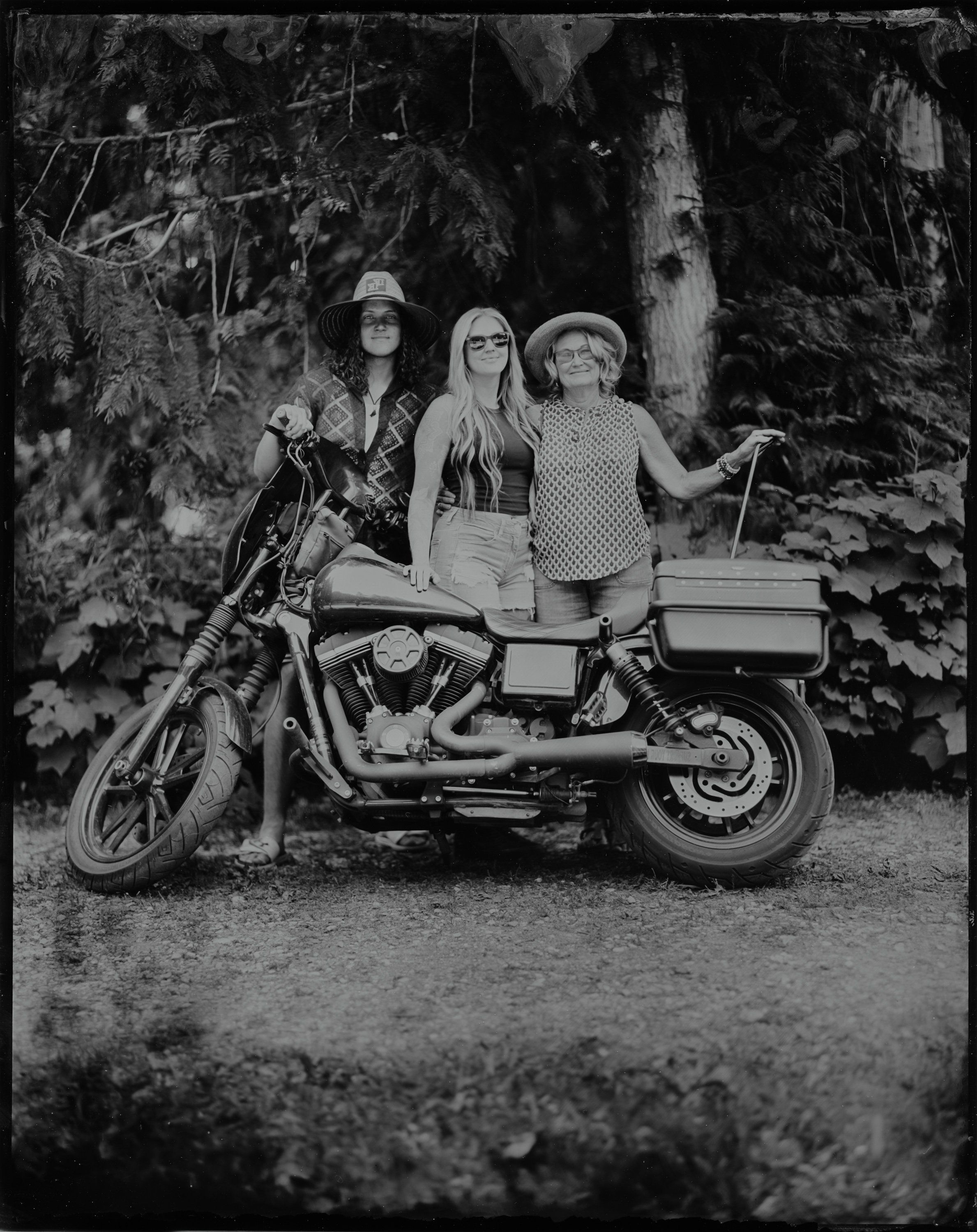 Three women pose on a vintage motorcycle with sidecar against tall trees, black and white photograph