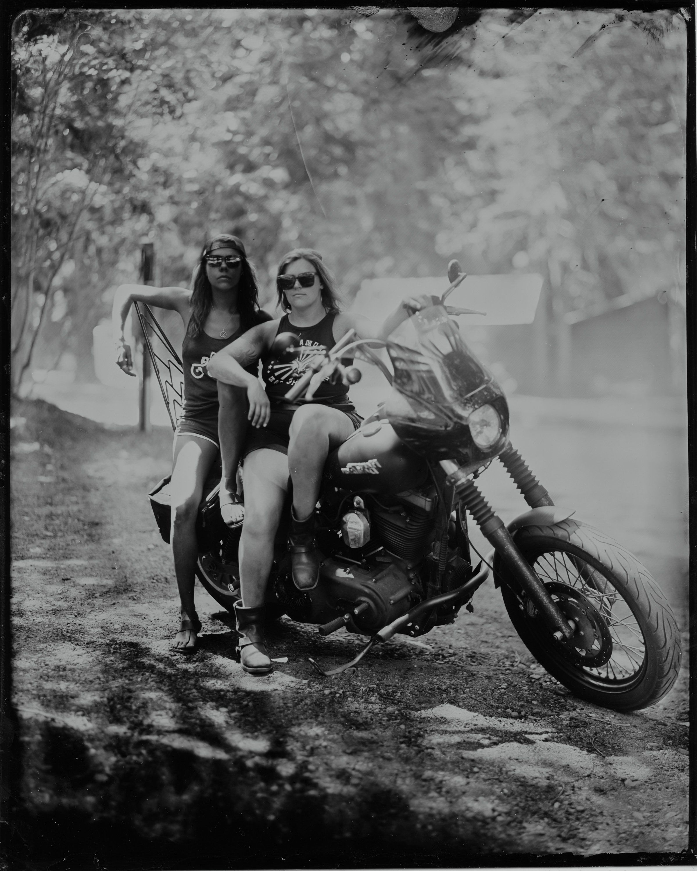 Two women pose on a vintage motorcycle on a tree-lined dirt road, dappled sunlight creating dramatic shadows across the scene