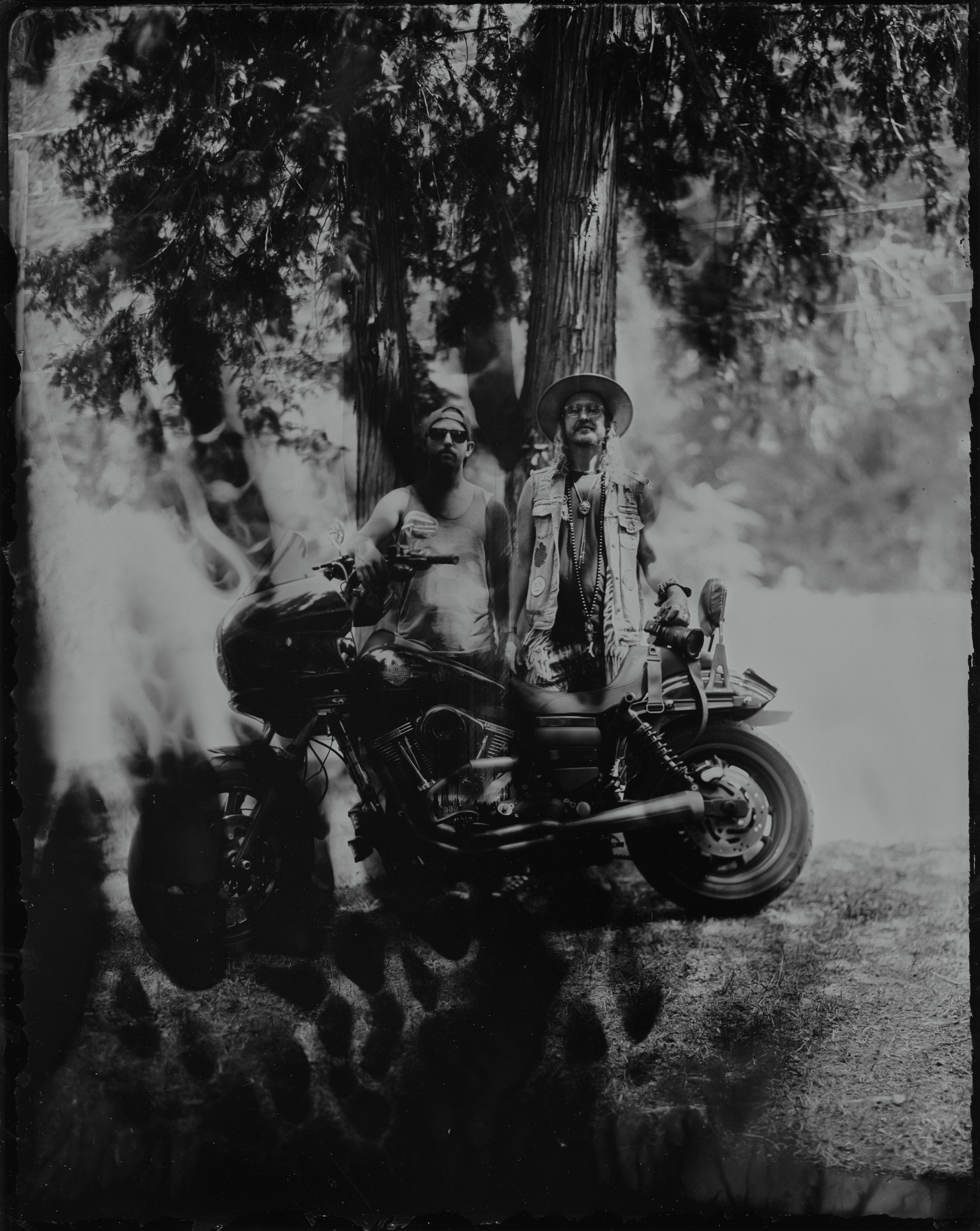 Two people on a motorcycle beneath a towering tree, dappled sunlight filtering through leaves in moody black and white