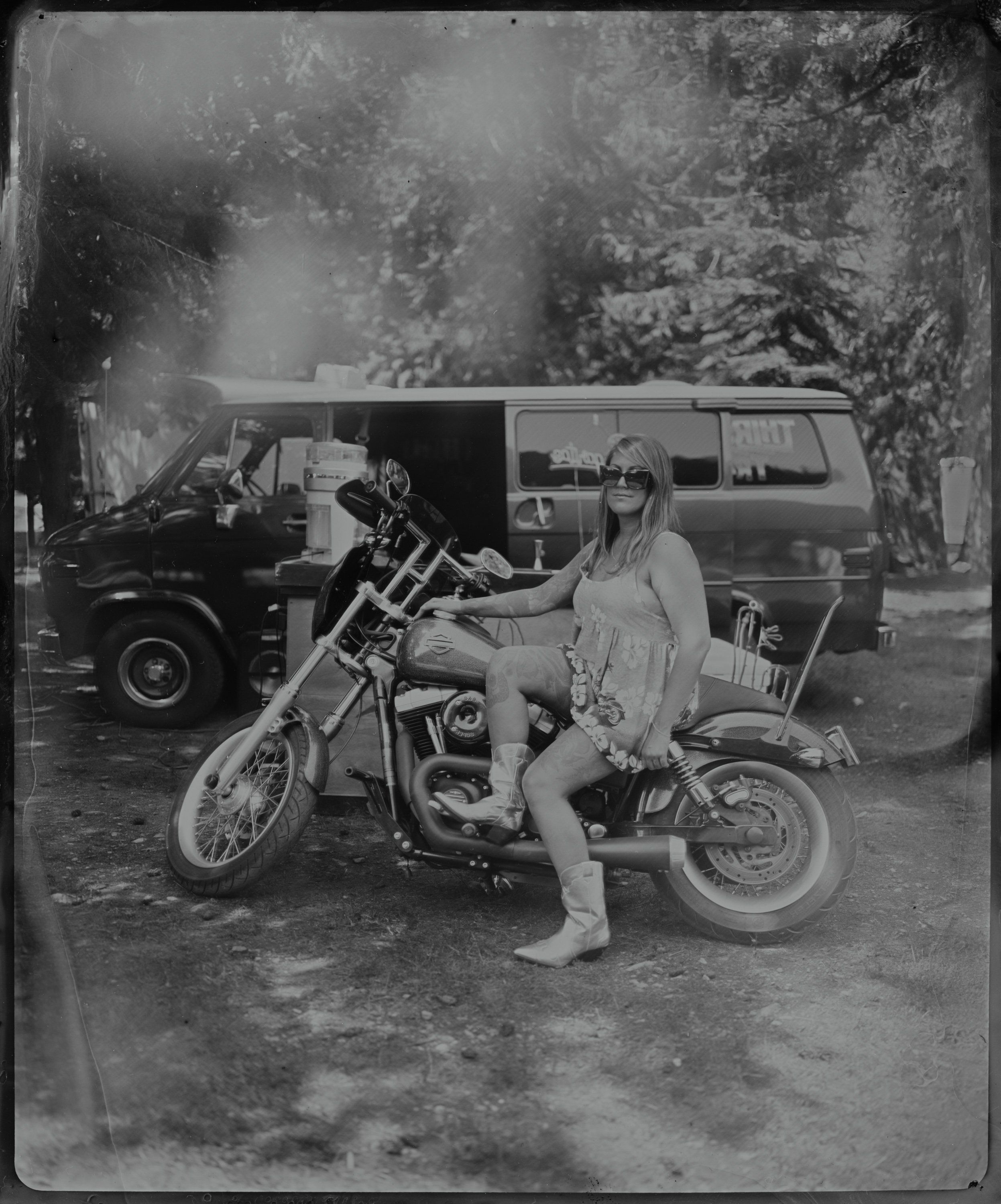 Woman in light dress seated on vintage motorcycle with chrome details, vintage van and trees in soft-focus background