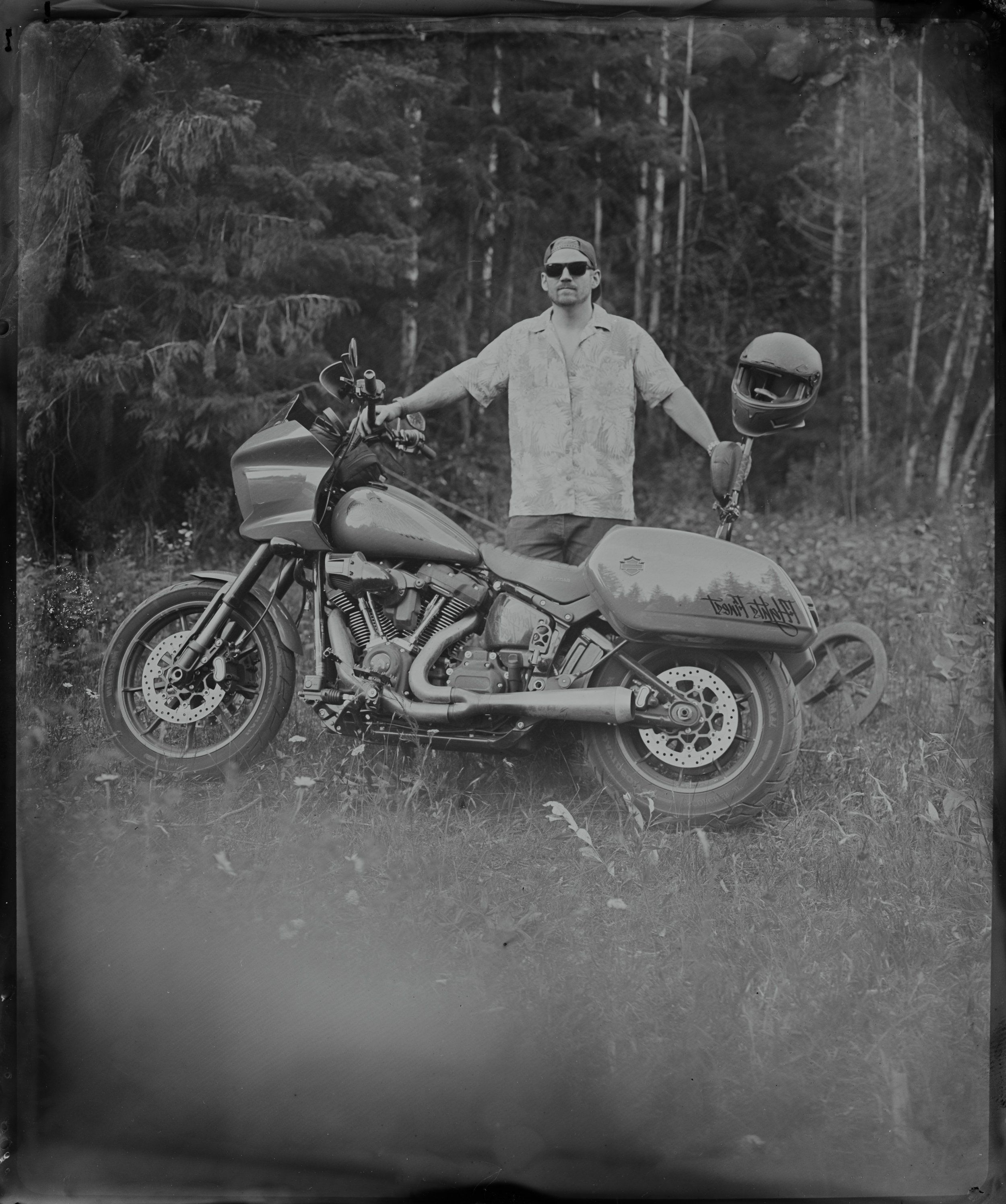 Man in sunglasses stands beside vintage motorcycle with sidecar in forested setting, holding helmet