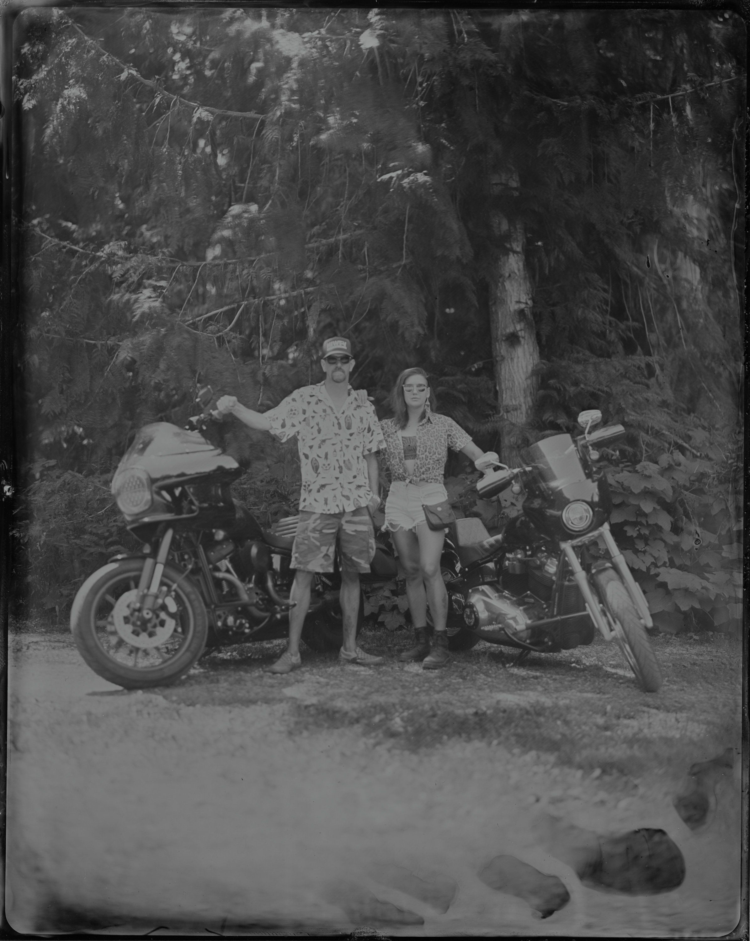 Two people stand with vintage motorcycles beneath towering forest trees in this grainy black and white photograph