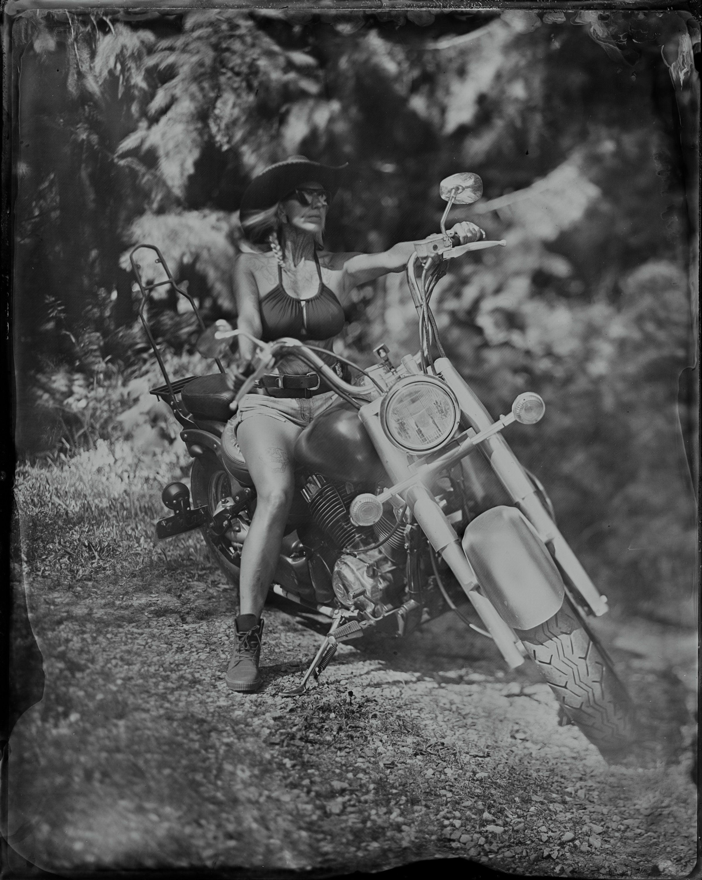 Woman in vintage dress and hat perched on classic motorcycle, arm extended in playful gesture amid dappled forest light