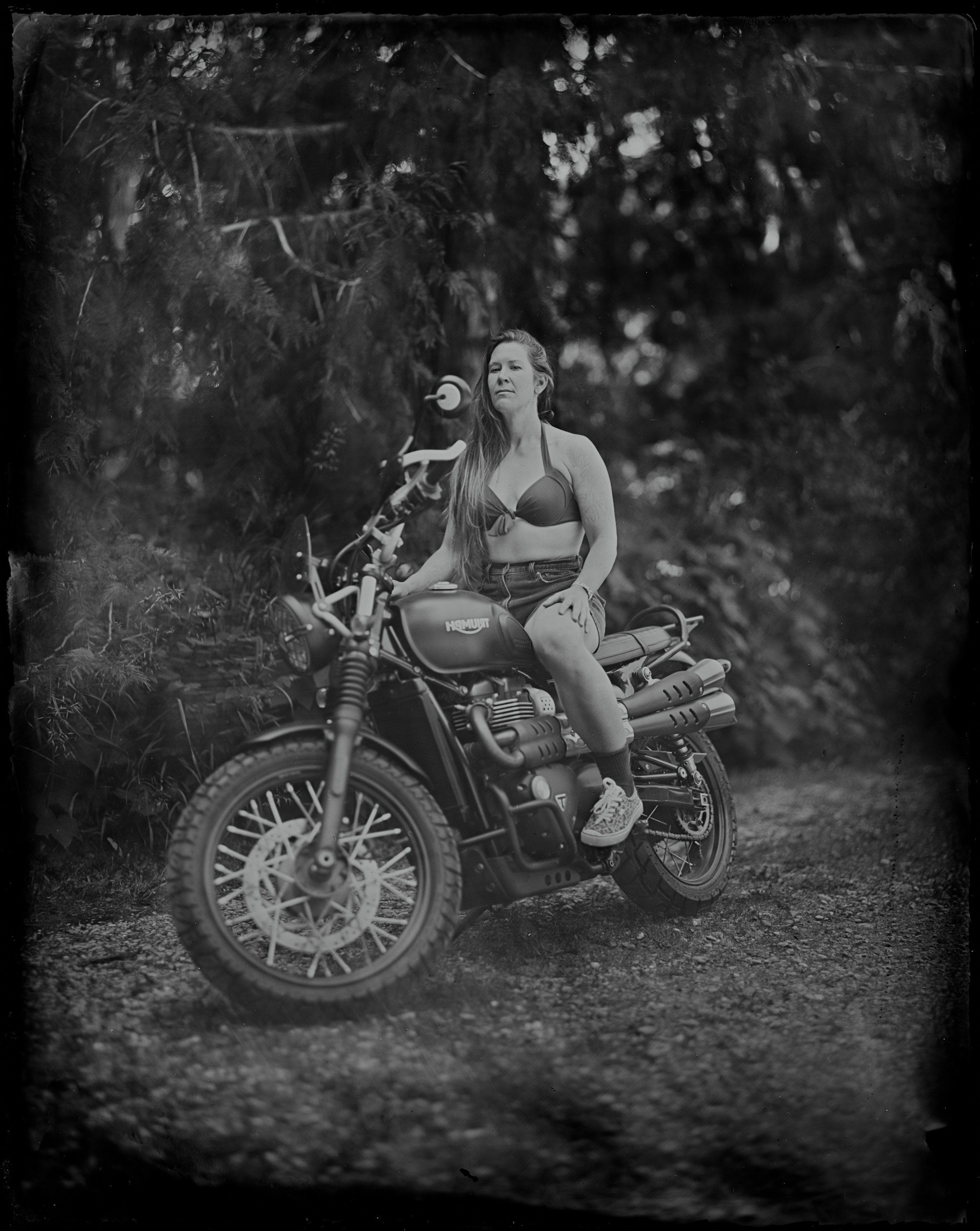 Woman in vintage bikini seated on classic motorcycle beneath dappled forest canopy, black and white portrait