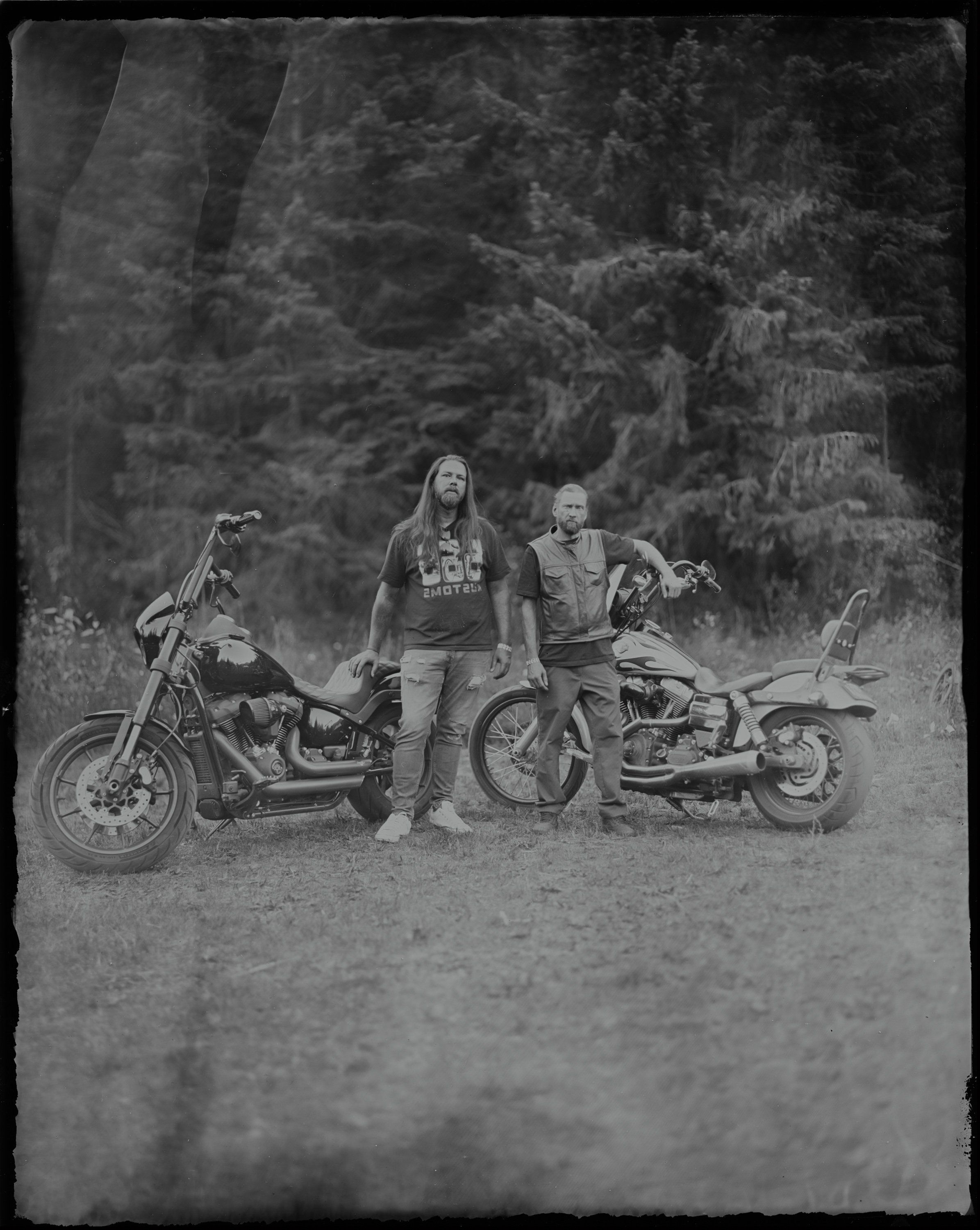 Two people with long hair stand between two custom motorcycles on a forest road, framed by dense evergreen trees in soft natural light