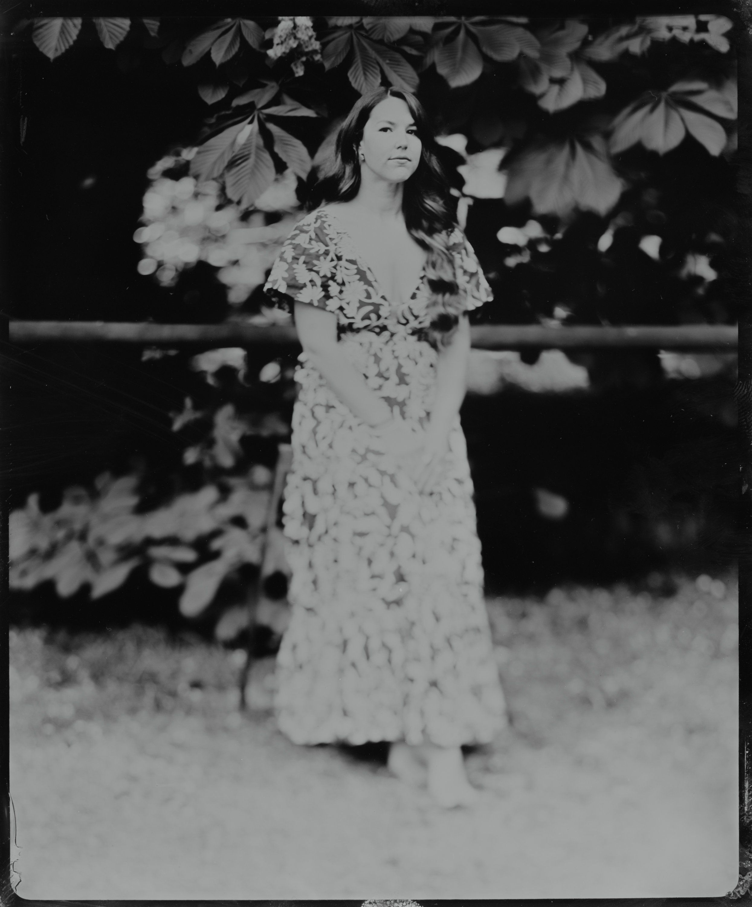 Woman in floral dress gazes upward beneath flowering branches, black and white portrait with soft natural light