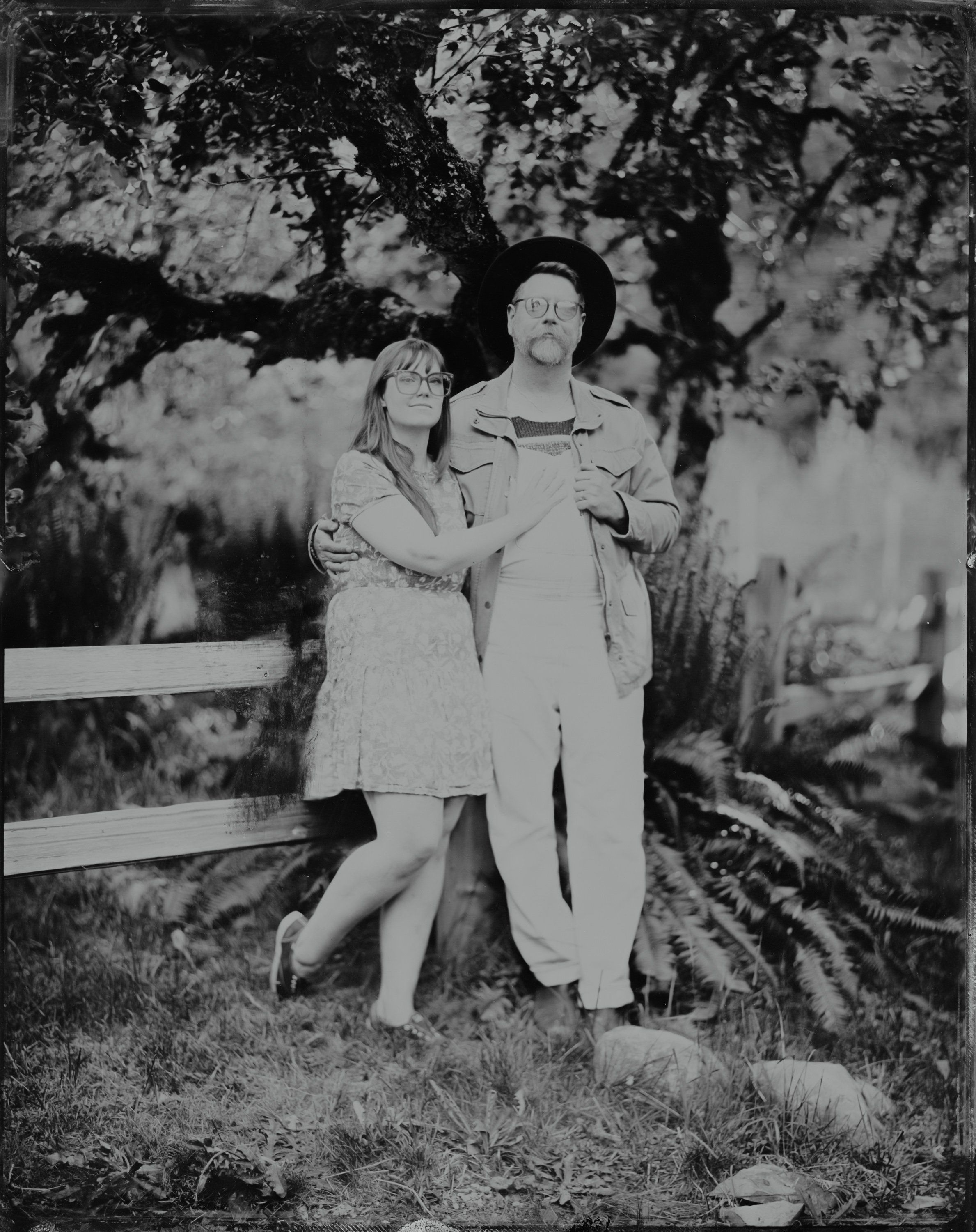 Couple in vintage attire standing beneath gnarled tree branches, black and white portrait with soft natural light filtering through foliage