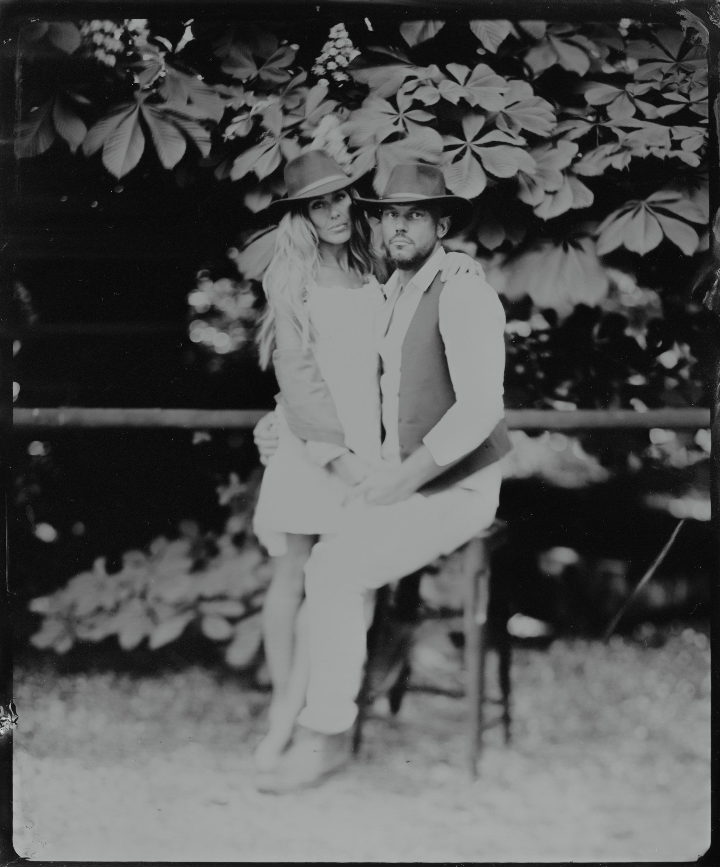 Man in light suit and hat seated beneath flowering vines, black and white portrait with soft natural light