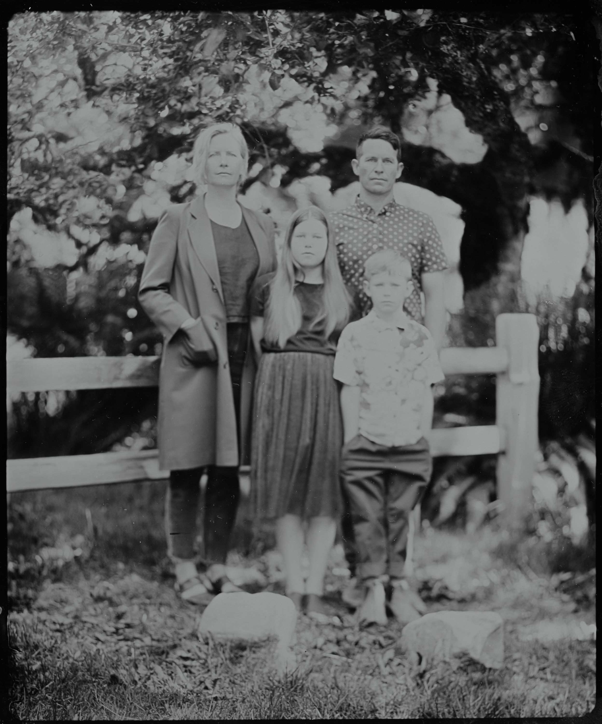 Three generations stand beneath dappled tree shade, their patterned clothing rendered soft in black and white film grain